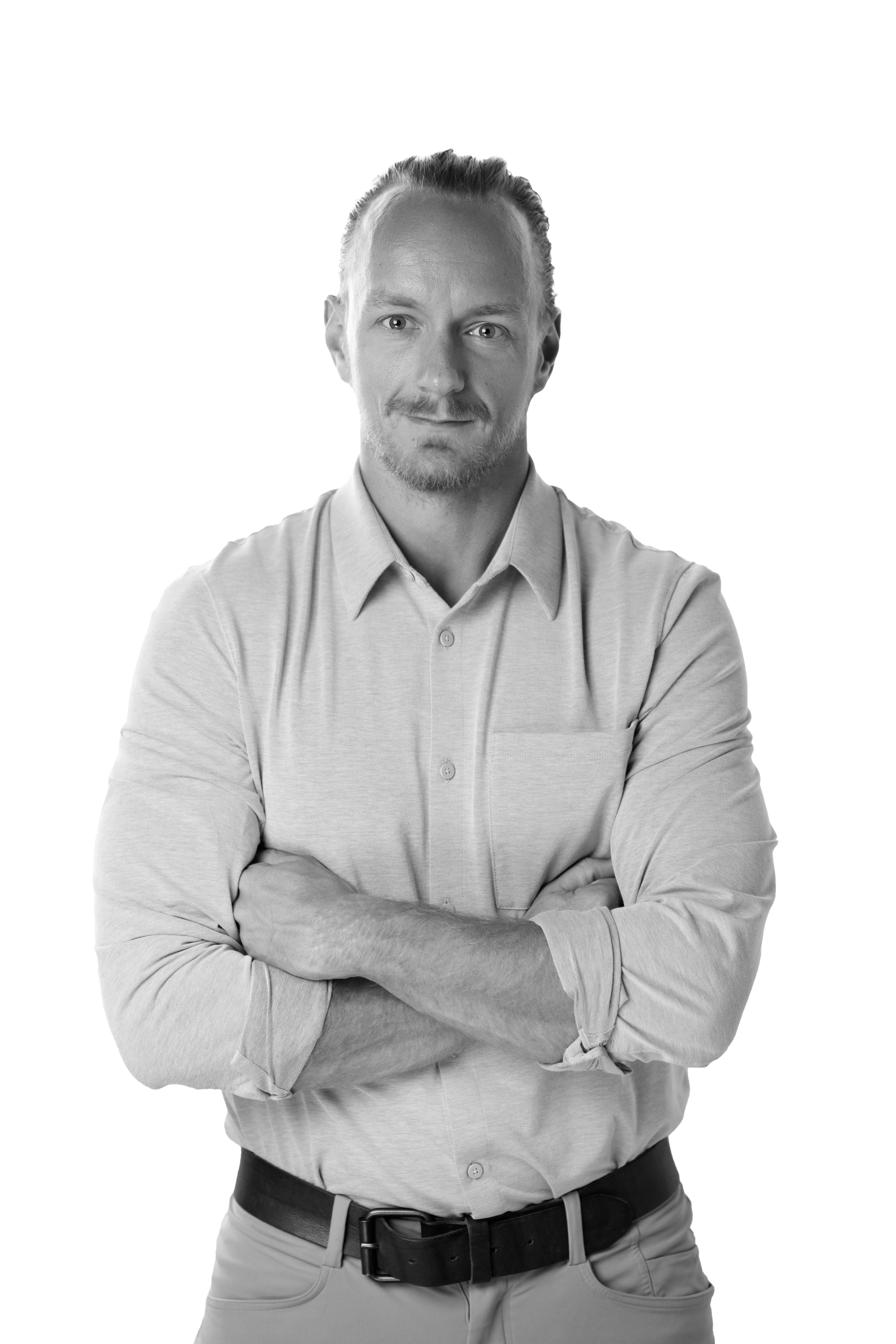 A man with short hair and a beard, wearing a light-colored dress shirt, crossing his arms, looking at the camera in a black-and-white portrait.