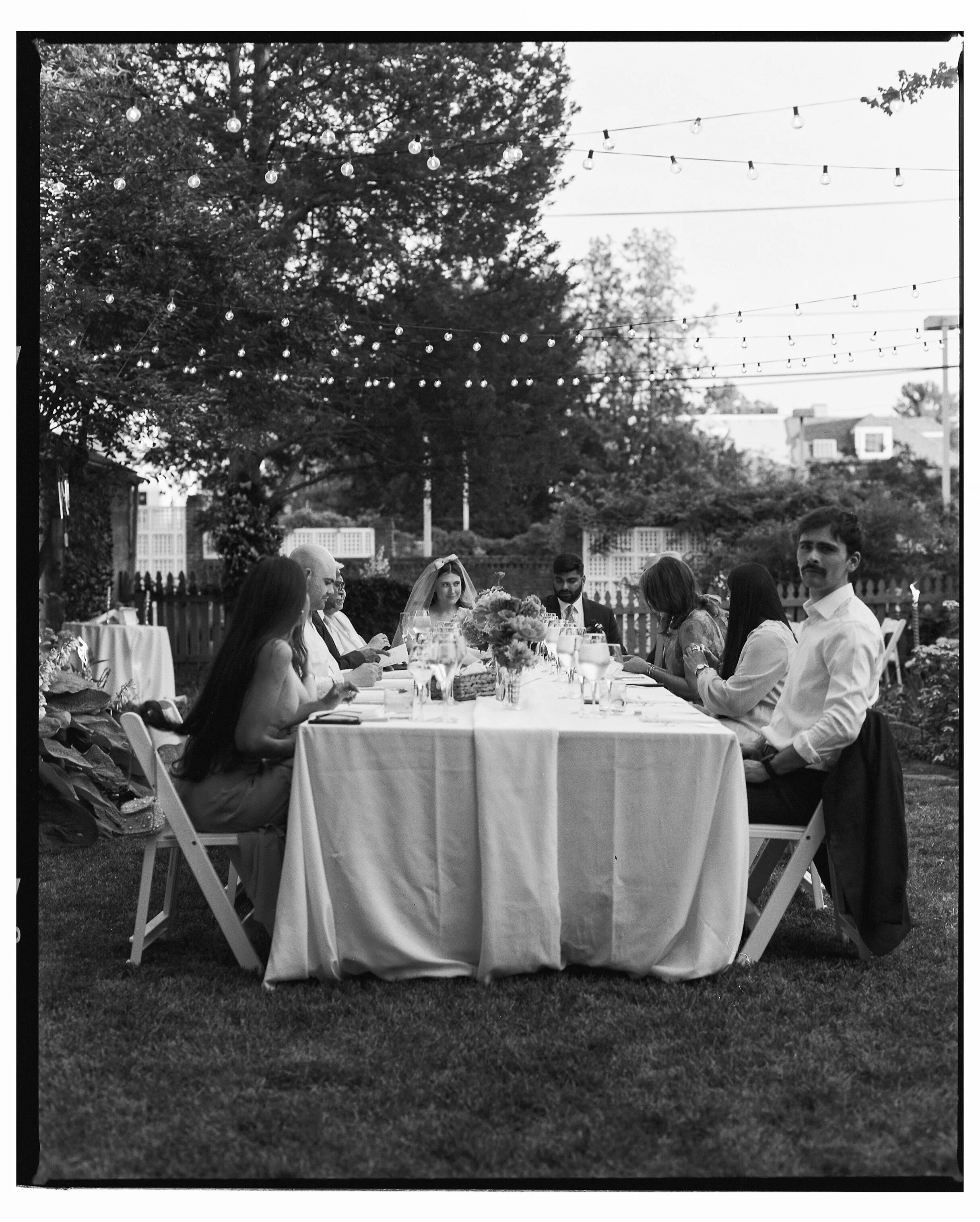 A black-and-white photo shows a small group of people seated around a round, cloth-covered table outdoors in a backyard setting. String lights are hung overhead, creating a soft, warm glow. The group is celebrating a garden wedding, with plates, glas