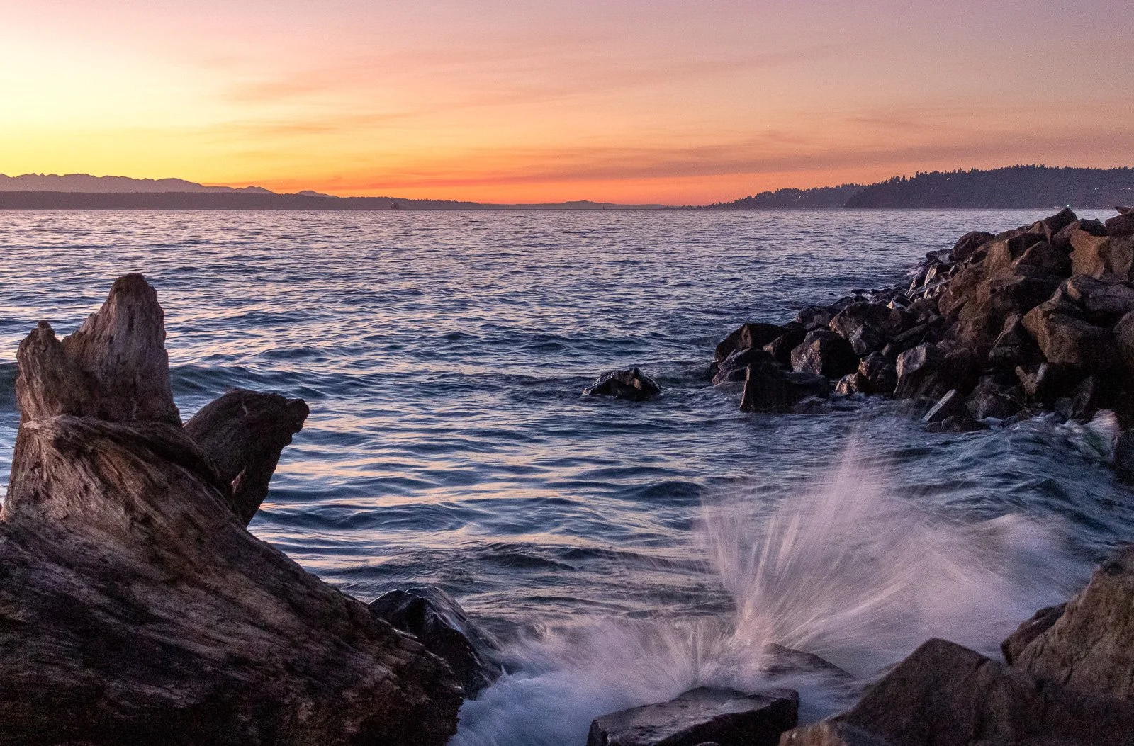Waves crash against rocky jetty boulders beneath a vivid orange and pink sunset along the Puget Sound shoreline