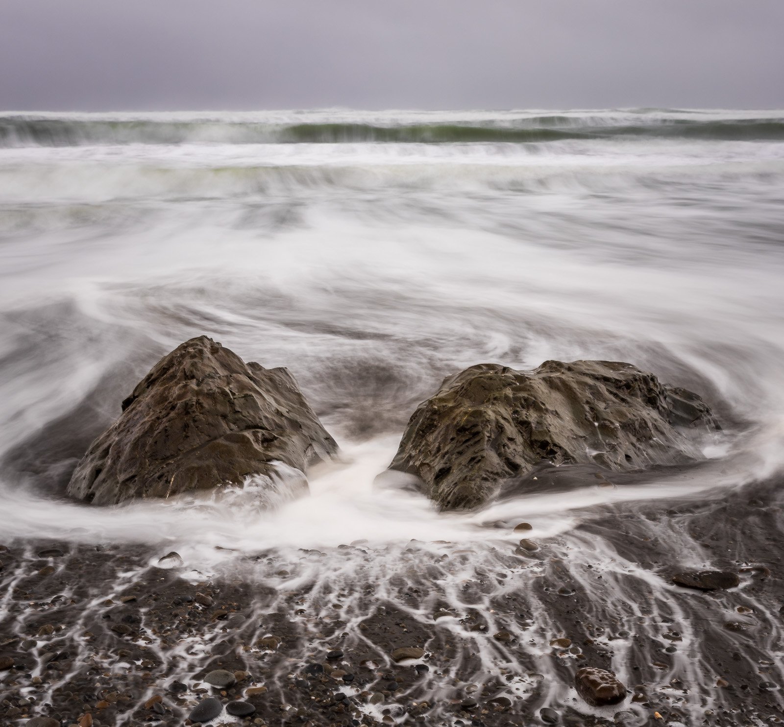 Long-exposure waves swirl around two exposed rocks on a moody gray morning along the Pacific Northwest shoreline