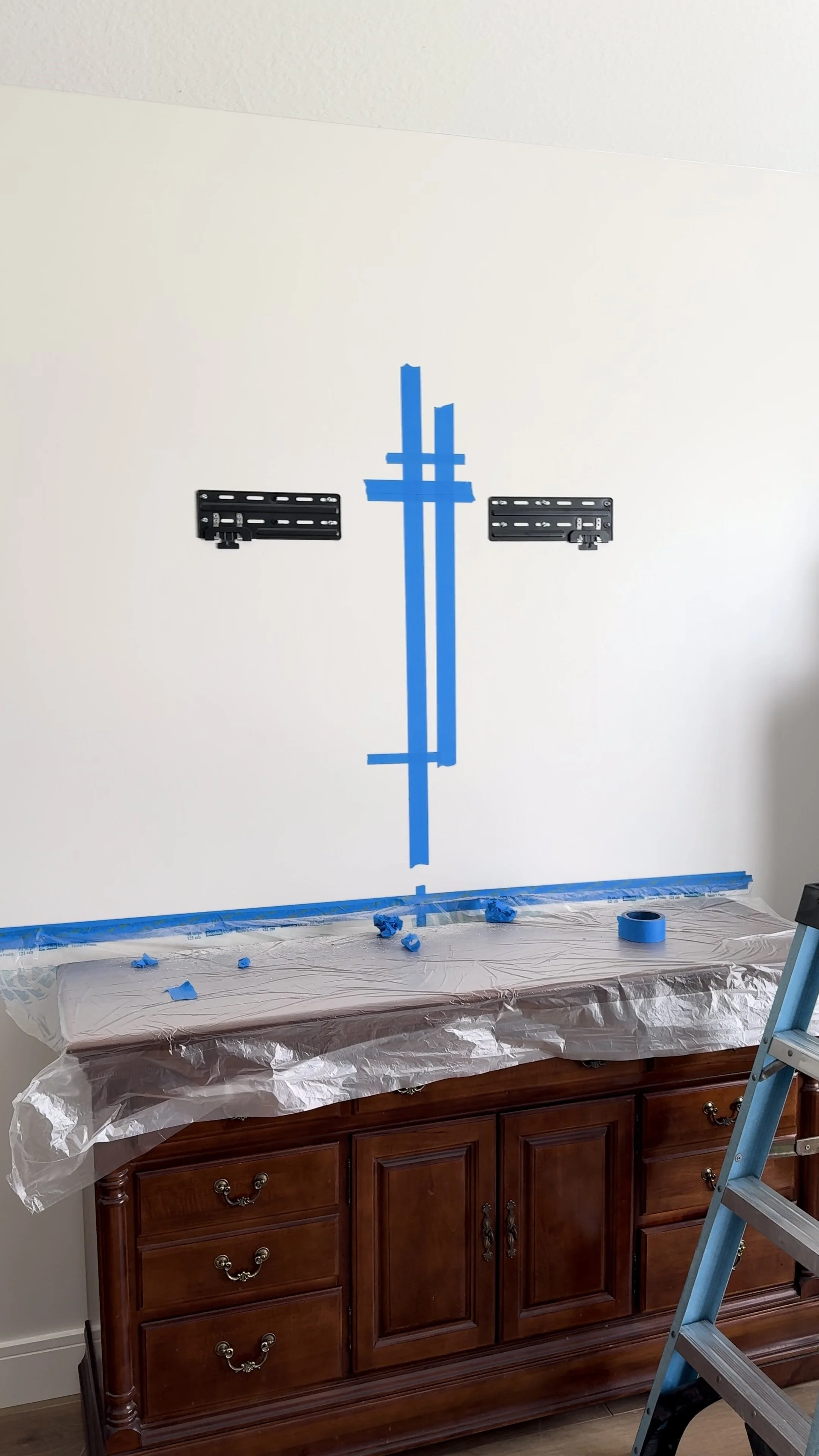A wall with mounting brackets and painter's tape arranged for picture frame installation, with a wooden cabinet below and a ladder nearby, in a room under renovation.