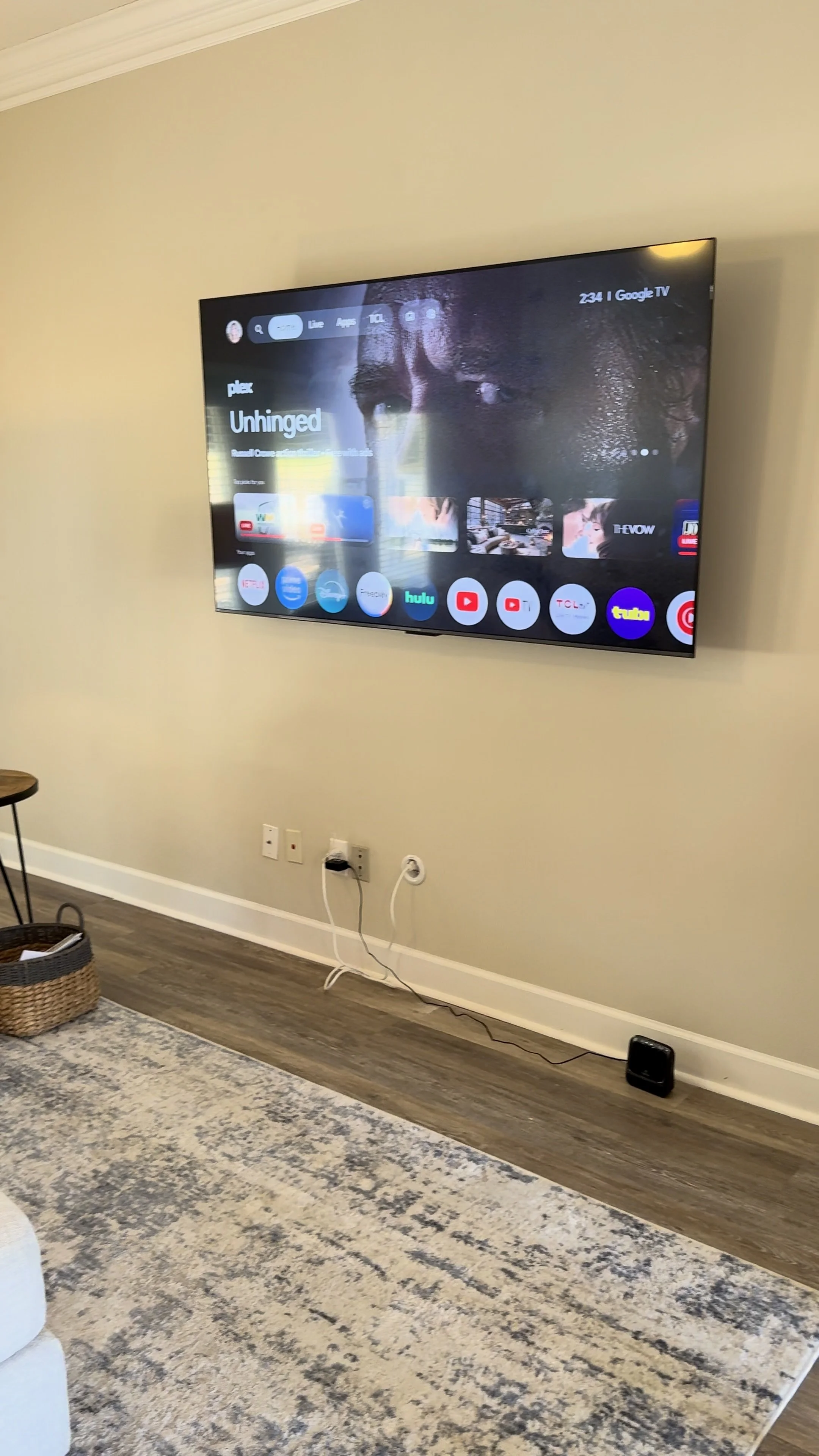 Living room with a wall-mounted television showing a menu screen with app icons, a beige wall, a patterned area rug, a small woven basket, and a small table