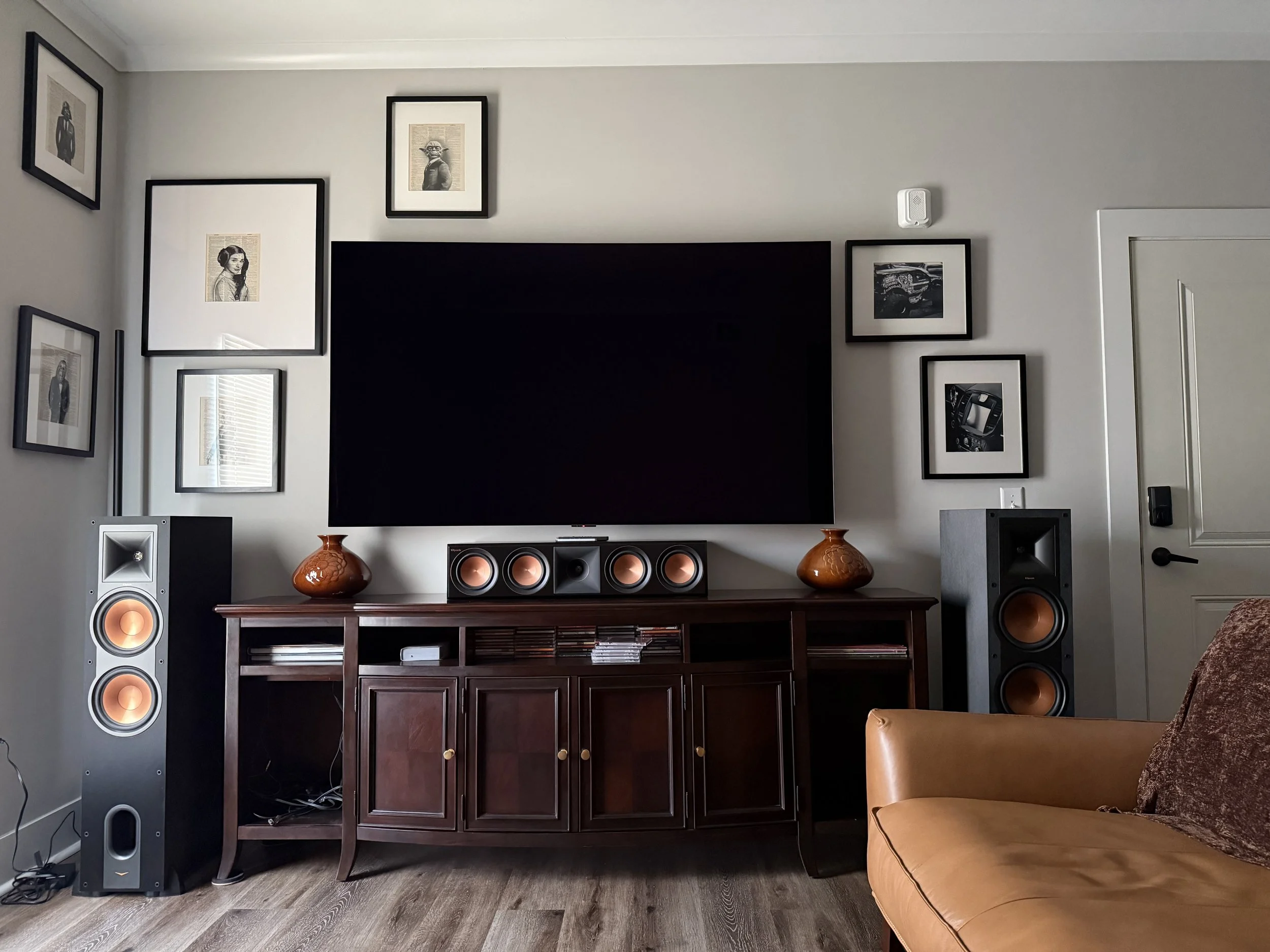 Living room with a large flat-screen TV, framed art on the wall, a wooden media console, a set of black and copper speakers, decorative vases, a leather armchair, and a section of a brown sofa.
