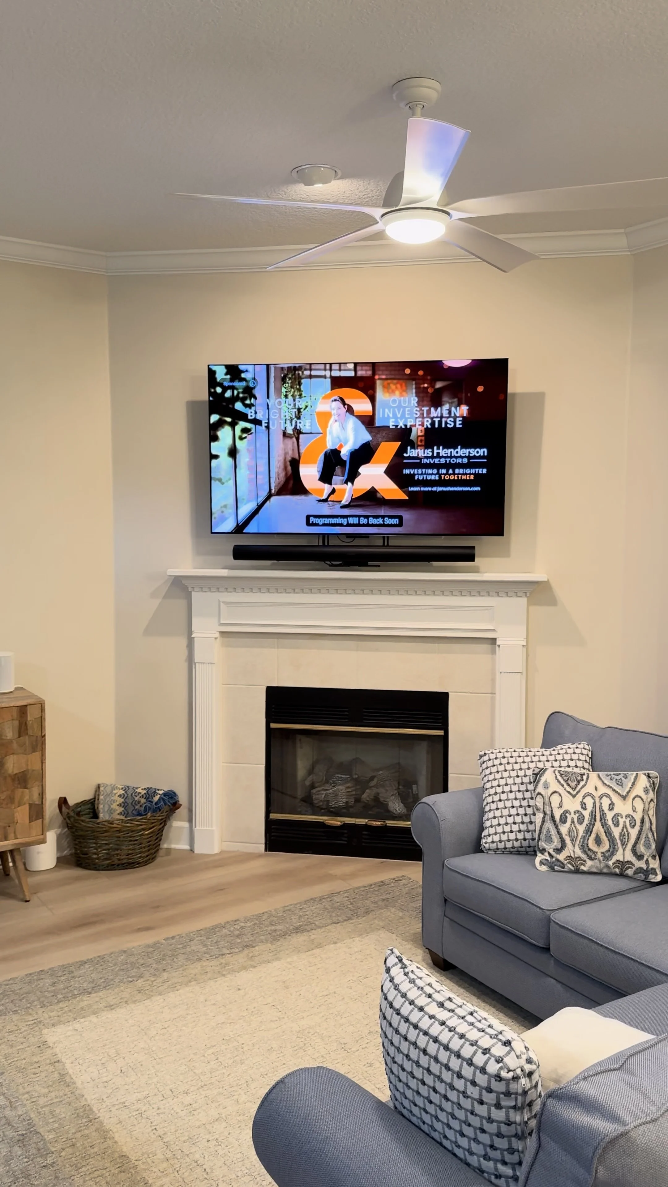A living room with a fireplace, a wall-mounted flat-screen TV displaying an investment advertisement, a ceiling fan with a light, a blue armchair with a patterned pillow, and a cream-colored area rug.