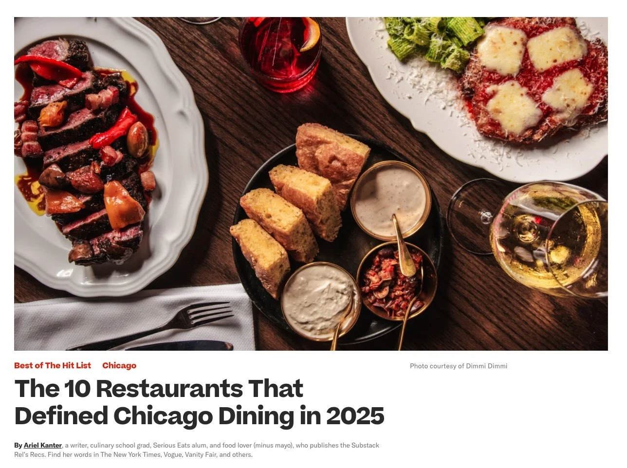 A top-down view of a dinner table with various dishes, including a plate of sliced steak with sauce, a pizza with cheese and greens, garlic bread, salads, and glasses of wine. A napkin with a fork and knife is also visible.