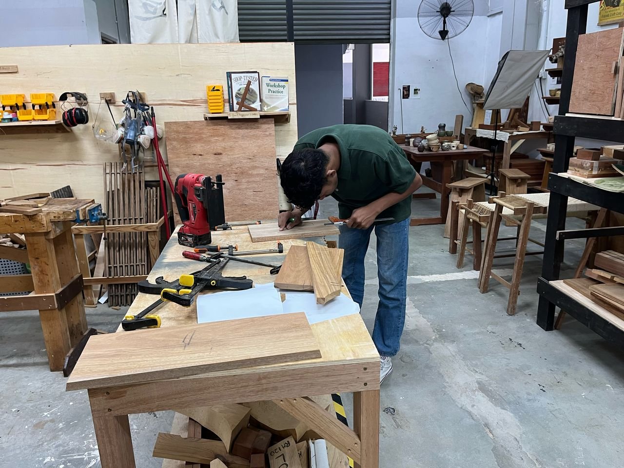 A person working in a woodworking workshop, using a ruler and pencil to measure a piece of wood on a workbench surrounded by various tools and wooden furniture pieces.