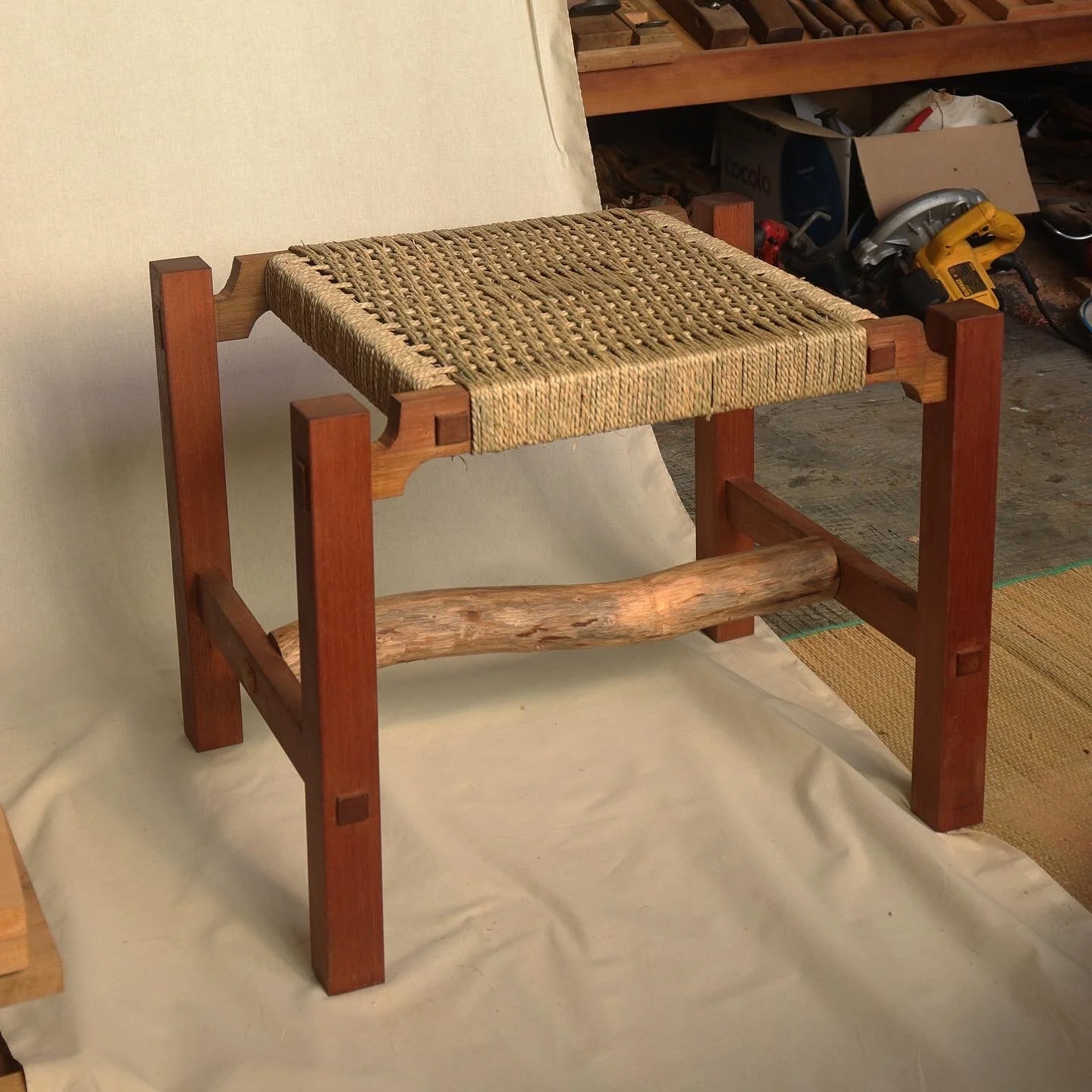 A wooden stool with a woven seat and a rustic wooden support beam underneath, placed on a neutral fabric background in a workshop setting.