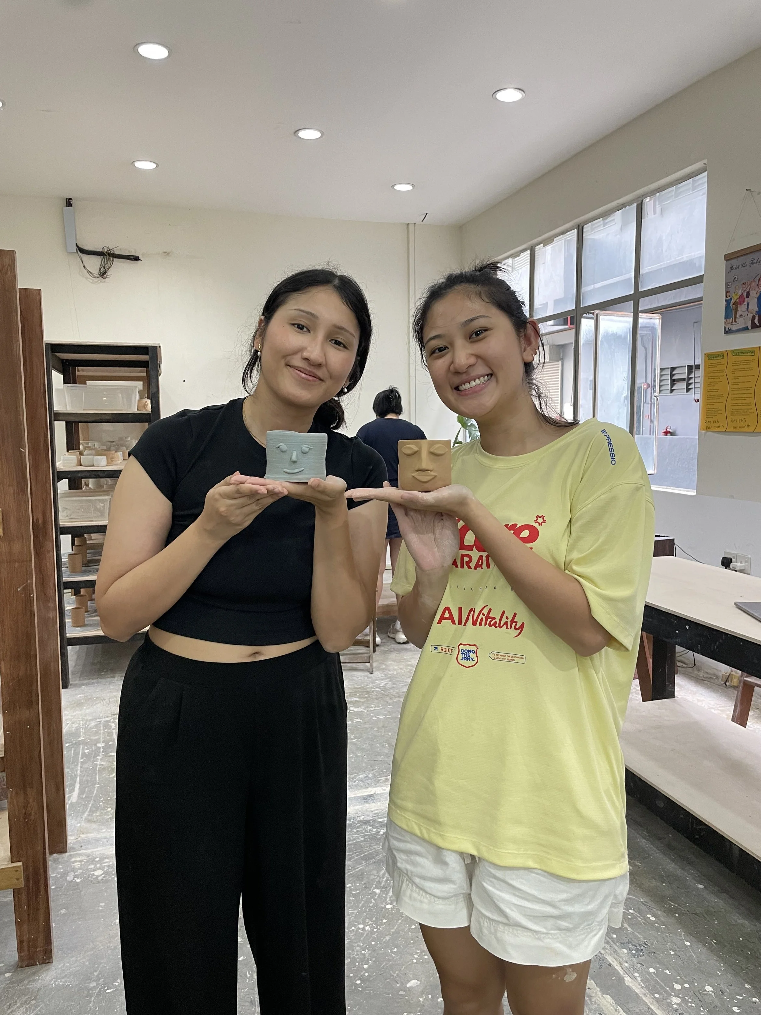 Two women smiling and holding small ceramic sculptures of faces in a pottery workshop at FRIENDS Craft Coop.