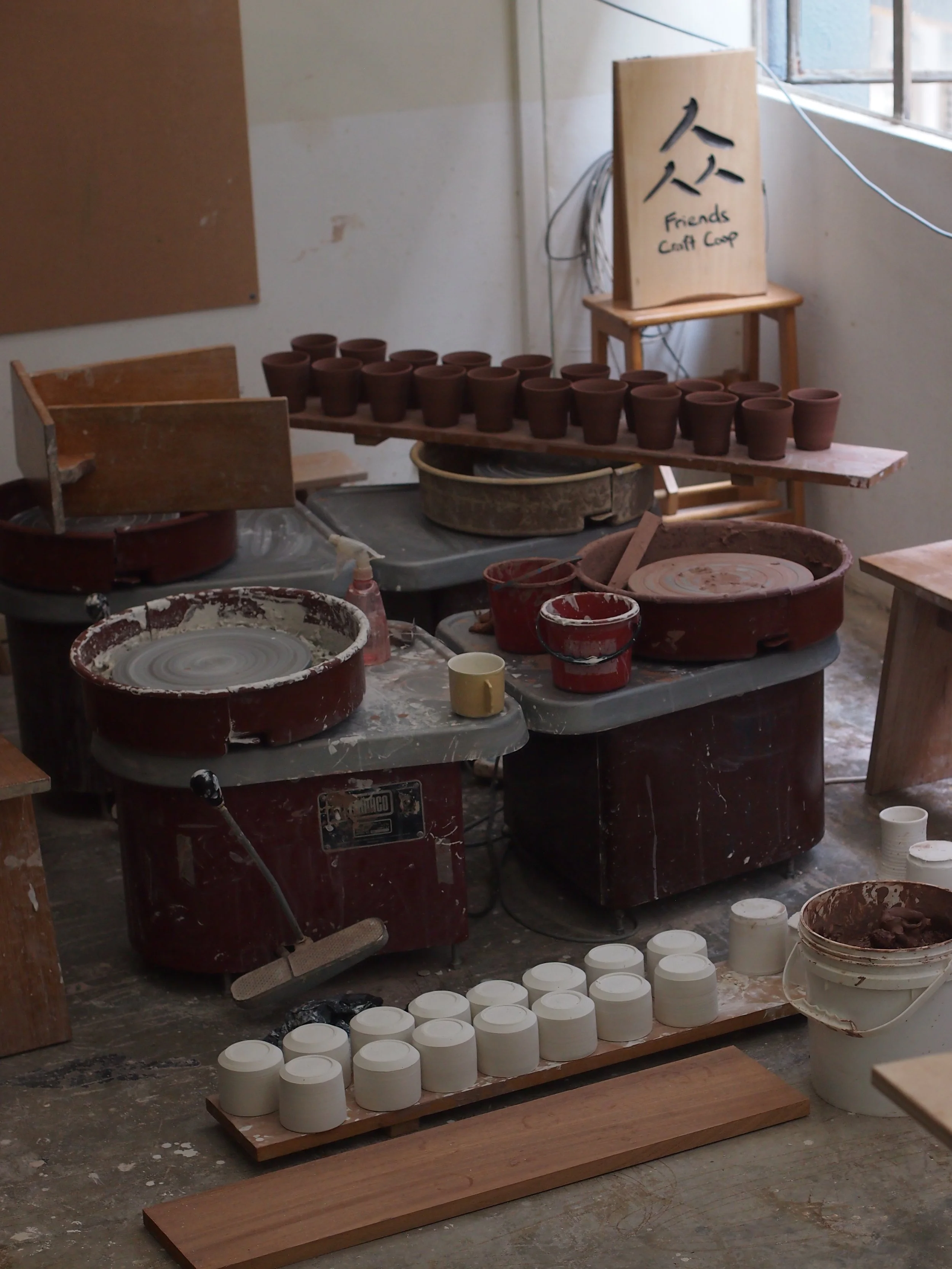 Freshly thrown unglazed clay cups on a wooden table, pottery wheels, and pottery tools scattered around, with a sign that says 'Friends Craft Coop' on a wooden stand near a window.