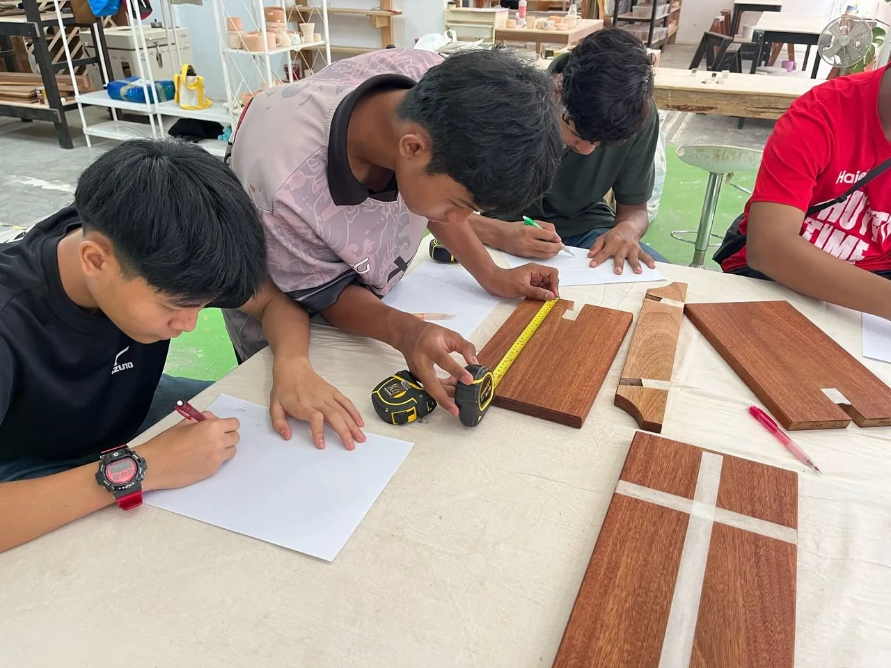 Four individuals working on woodworking or furniture project at a workshop, measuring and sketching while wooden pieces are laid out on the table.