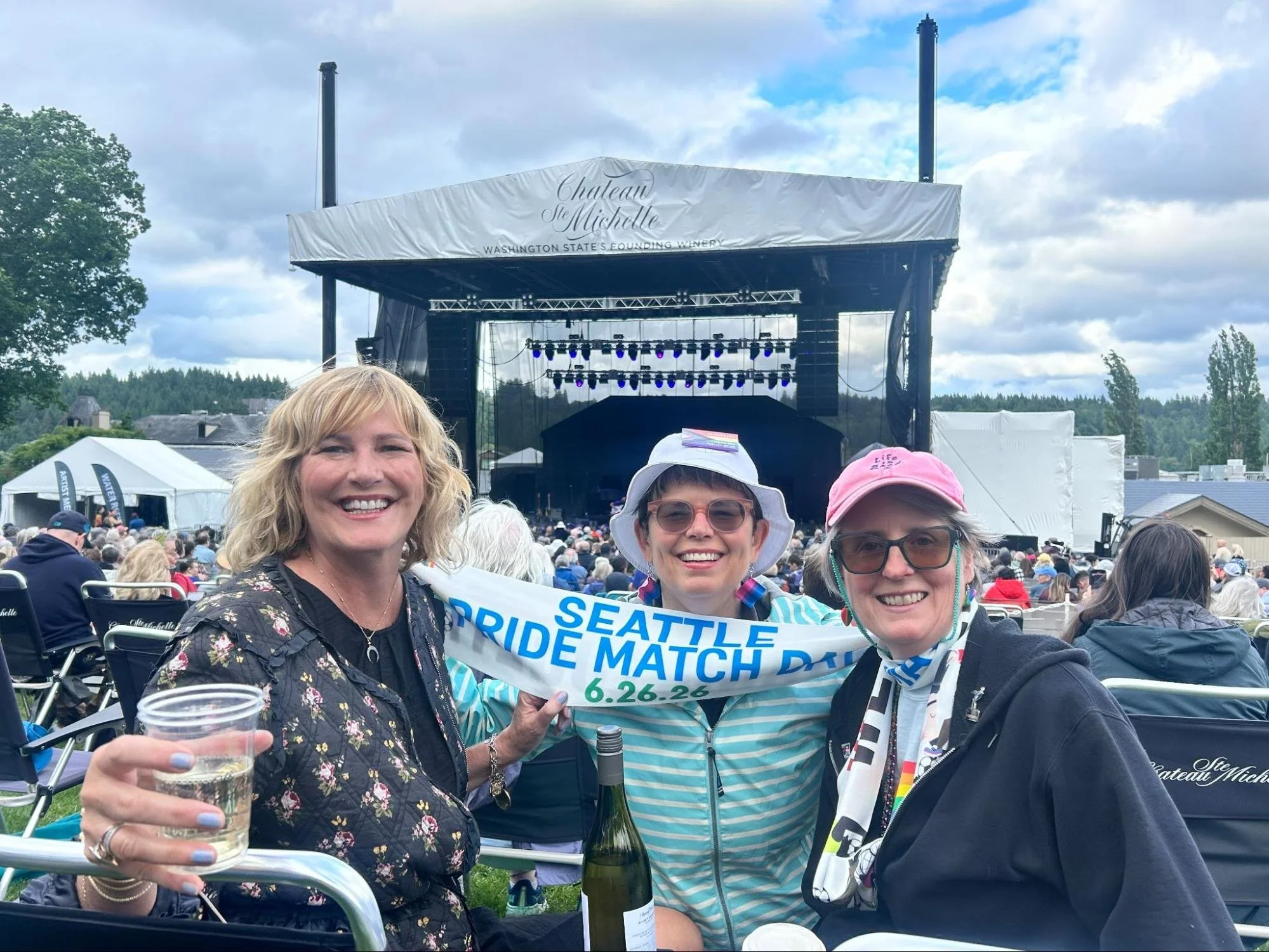 Three women smiling and holding drinks at an outdoor Pride march event, with a stage and lake visible in the background.
