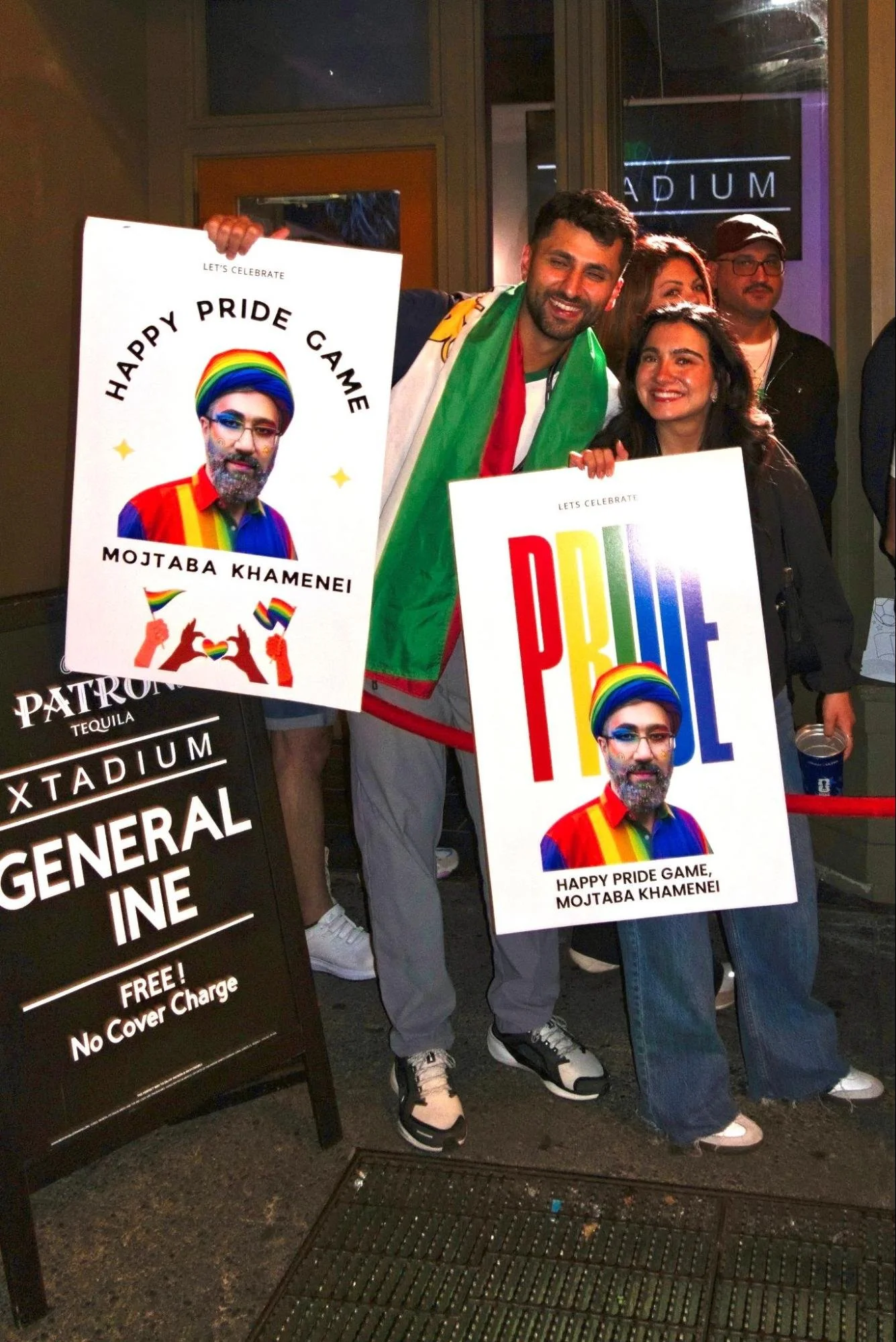 Two people outside a stadium entrance at night holding Pride-themed signs featuring an illustrated figure in a rainbow-striped shirt and beanie, one reading 'HAPPY PRIDE GAME, MOJTABA KHAMENEI' and the other 'PRIDE.'