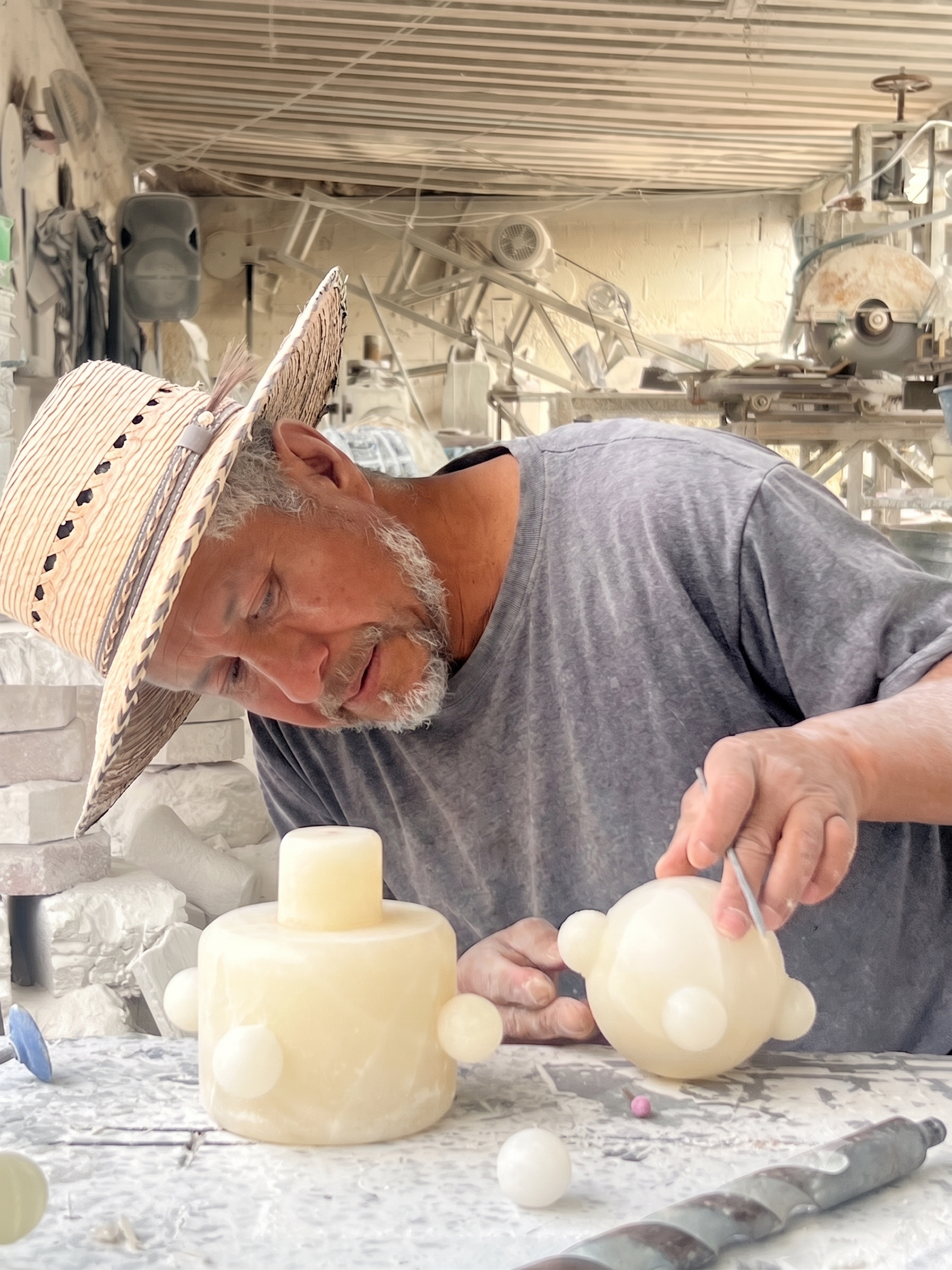 Hombre mayor trabajando en una escultura de cera en un taller, usando un sombrero de paja y ropa gris, con varias piezas de cera y herramientas en la mesa.