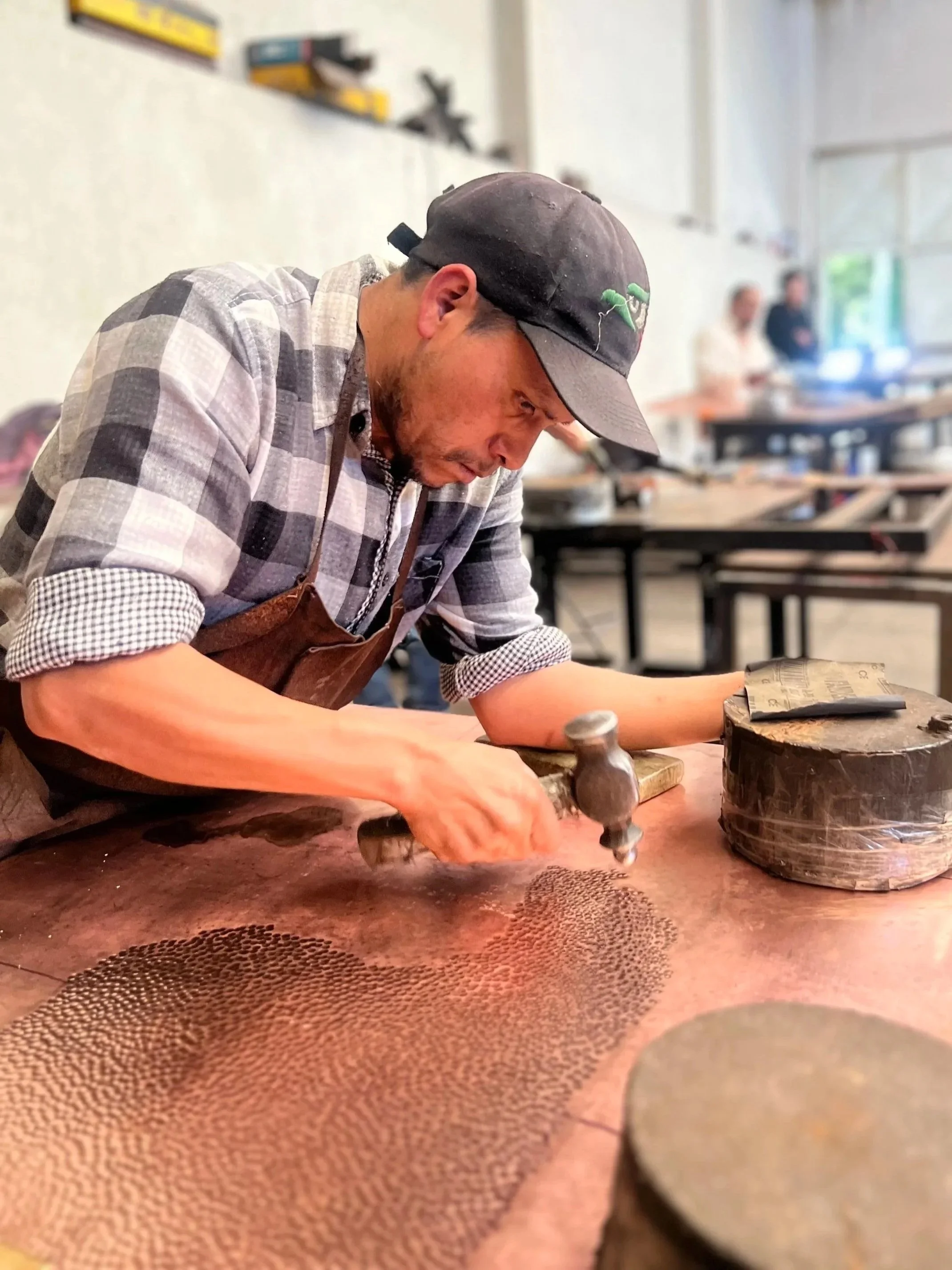 Un hombre con gorra y camisa de cuadros trabaja en un taller, concentrado en su tarea mientras manipula objetos pequeños sobre una mesa de trabajo.