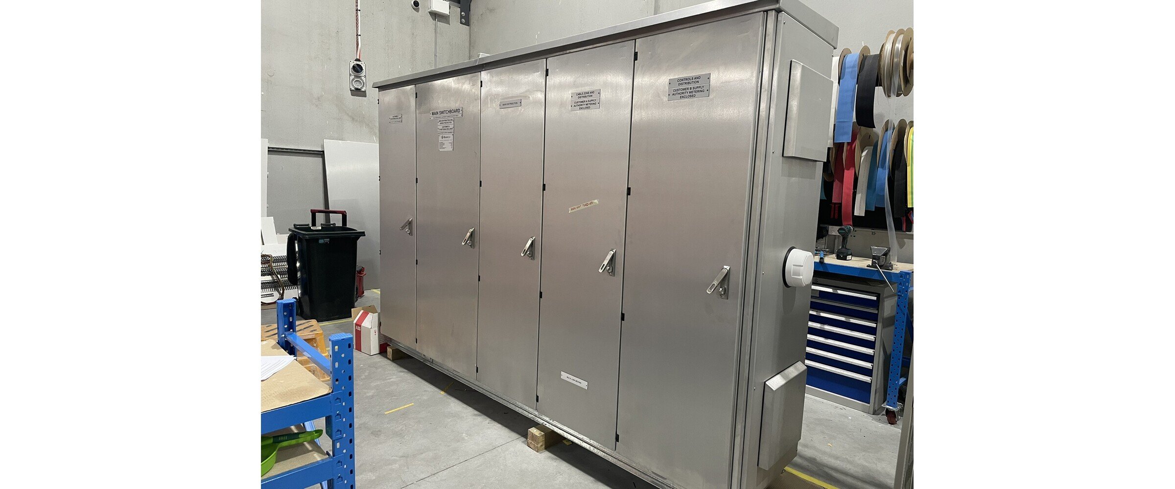 A large stainless steel electrical equipment cabinet with multiple doors and labels, situated in an industrial setting with a blue tool cart and various equipment nearby.