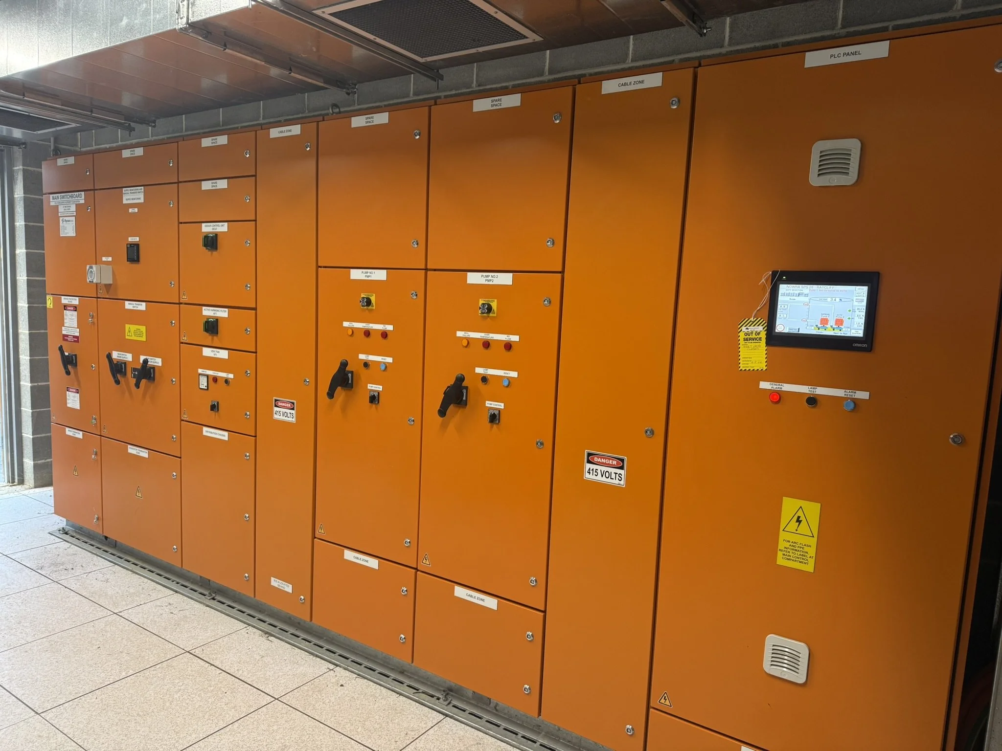 A large orange electrical control panel with various switches, labels, and warning signs, located indoors on a tiled floor.