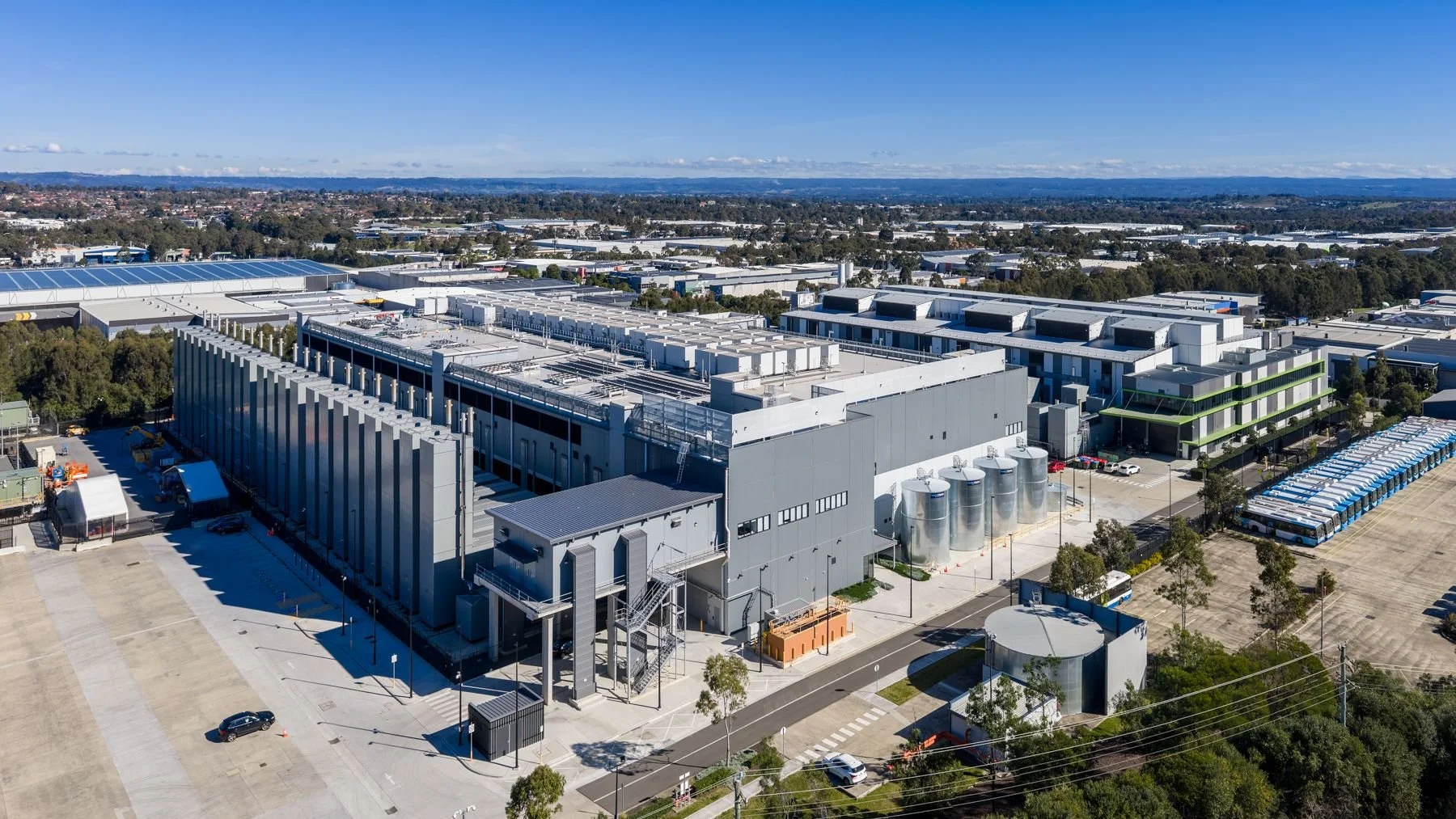 An aerial view of a large, modern industrial or technology facility with multiple flat roof buildings, silos, and parking lots, surrounded by trees and a vast landscape under a clear blue sky.