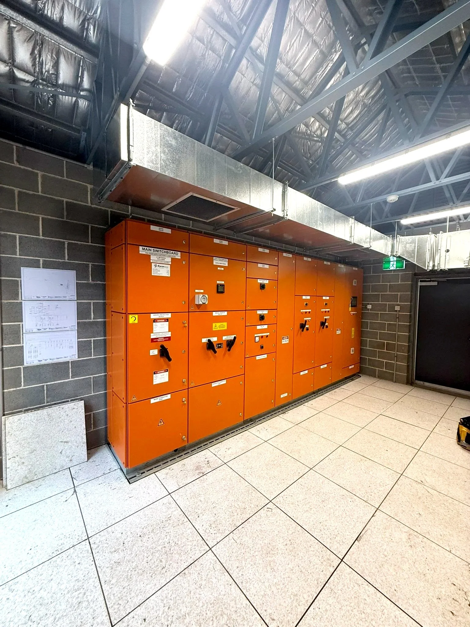Orange electrical control panels in a room with tiled floor and brick walls, with a large metal HVAC duct running overhead and fluorescent lighting on the ceiling.