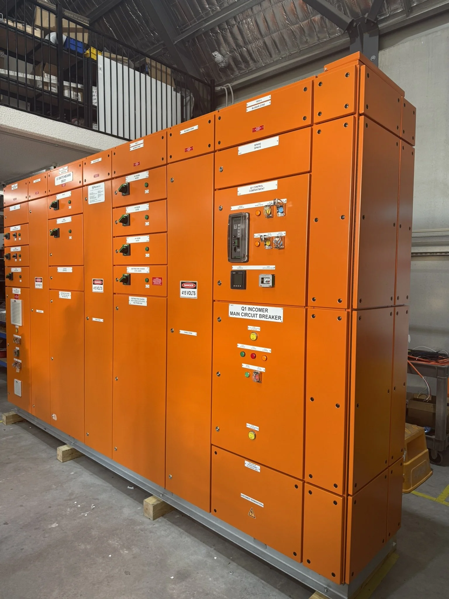 Orange electrical switchgear cabinet with labeled compartments and controls, resting on wooden blocks in an industrial setting.