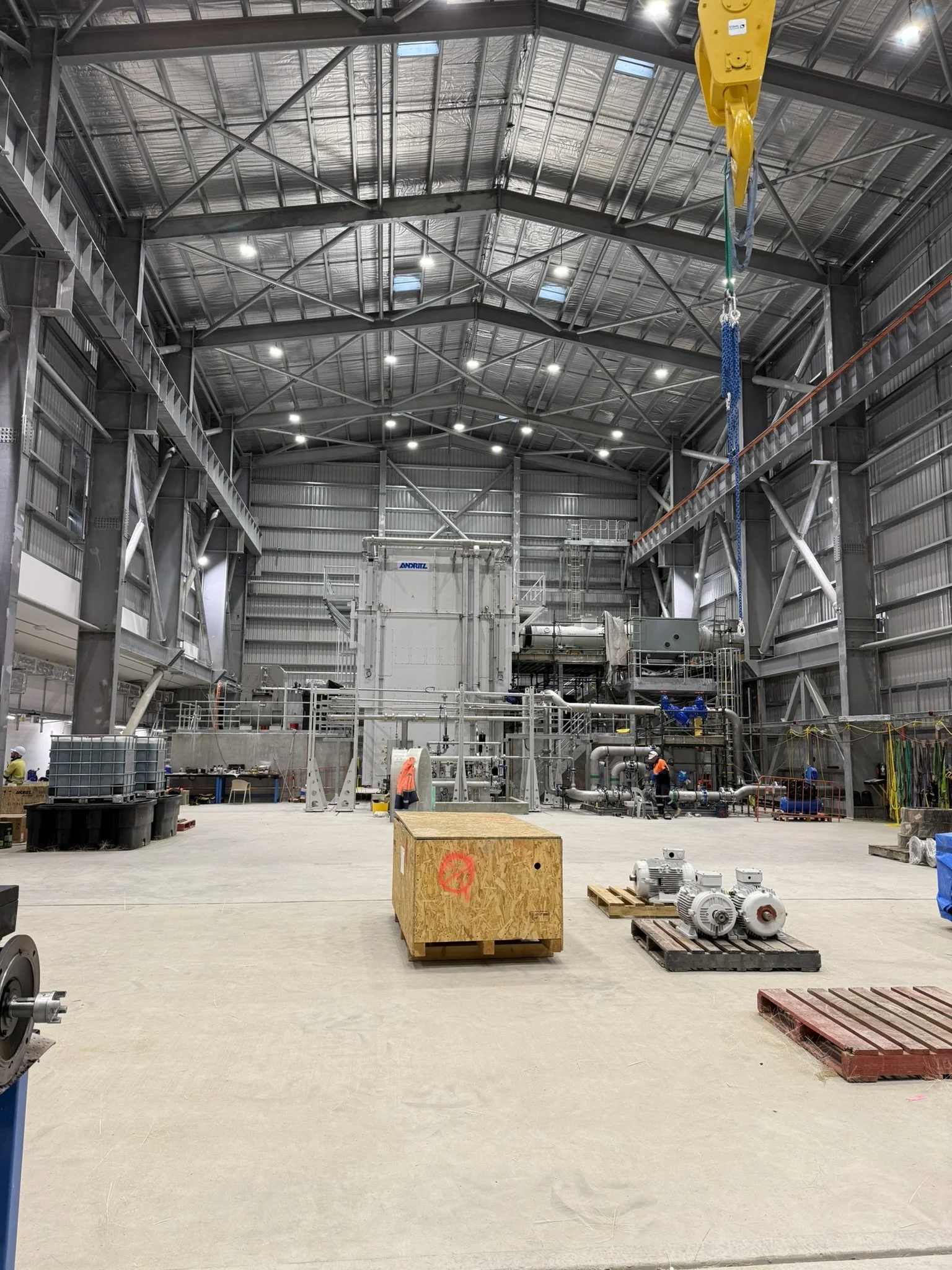 Inside a large industrial warehouse with steel beams and a high ceiling, workers in safety gear are working near machinery and equipment, including pipes, motors, and pallets.