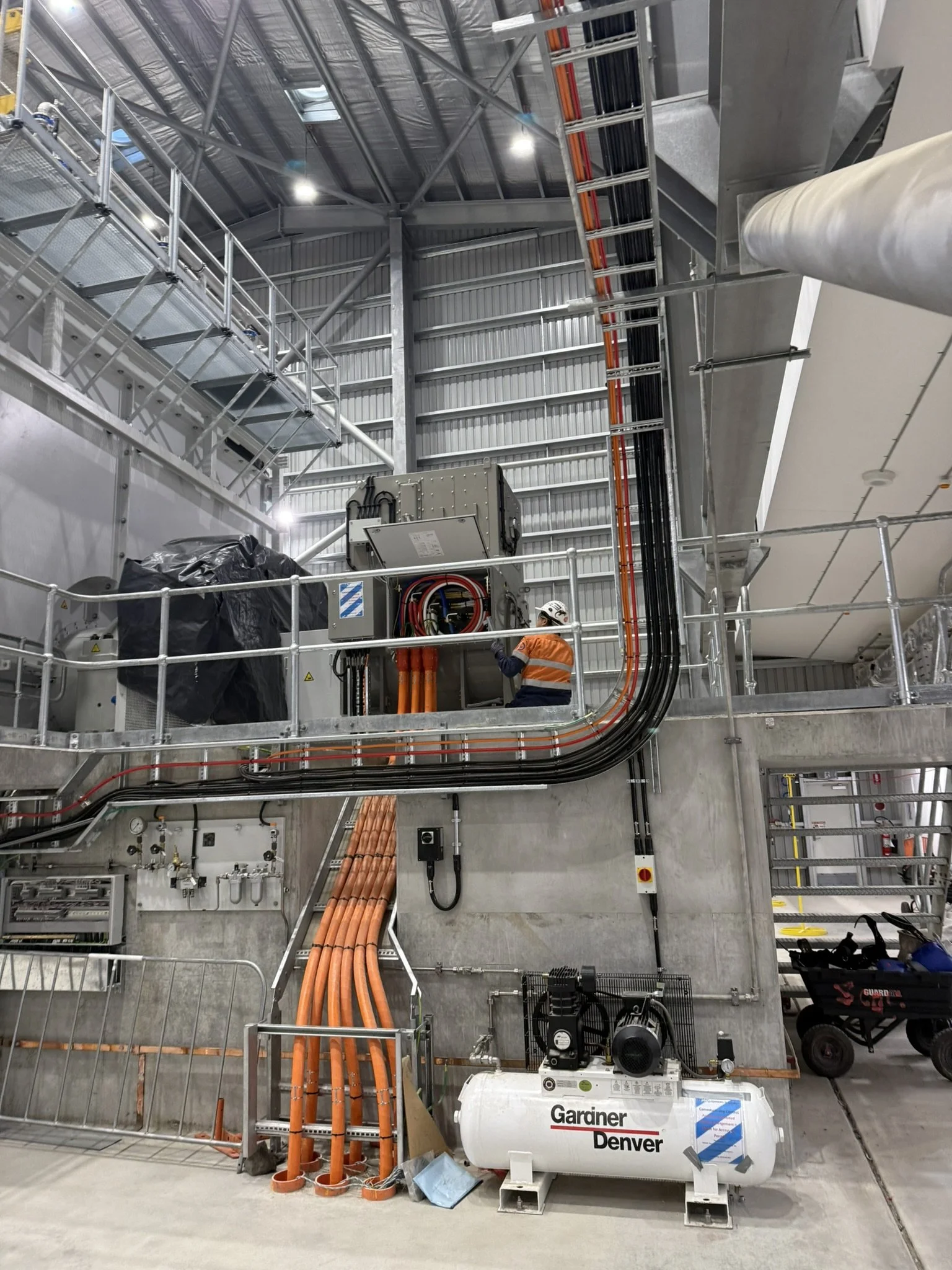 Construction site interior with steel framework, pipes, and electrical systems. Two construction workers in safety vests and helmets working near large equipment, including a white tank labeled 'Garaner Denver'.