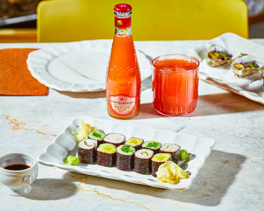 Suşi ruloları, beyaz bir tabakta zencefil ve wasabi ile servis ediliyor. Yanında kırmızı içecek bulunan bir şişe ve mermer masada bardak yer alıyor.

