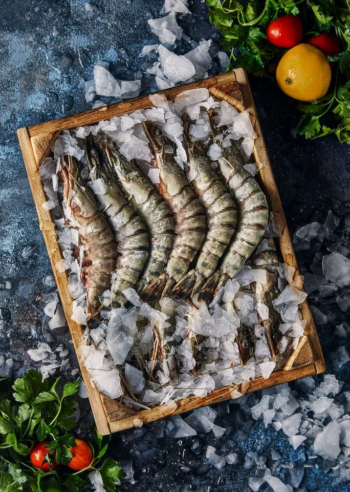 Maydanoz, domates ve limon ile çevrelenmiş, buz üzerinde çiğ kaplan karideslerinin yer aldığı ahşap tepsi