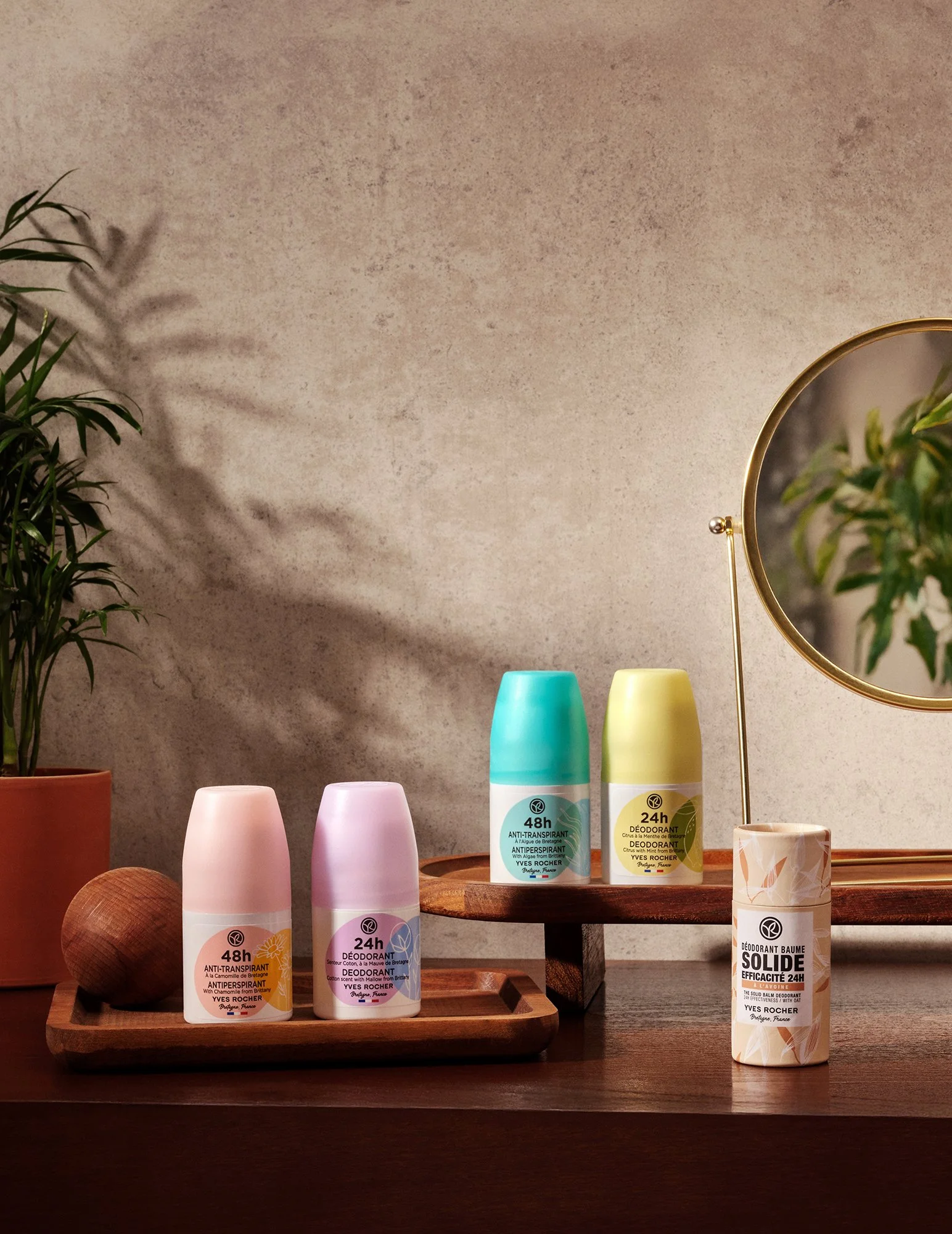 Pastel renk kapaklı beş Yves Rocher deodorant stick, ahşap tepsiler üzerinde sergilenmiş; yanında küçük bakır renkli bir top ve ahşap yüzeyde yuvarlak bir ayna bulunuyor. Arka planda saksılı bir bitki ve dokulu duvara düşen bir gölge var.