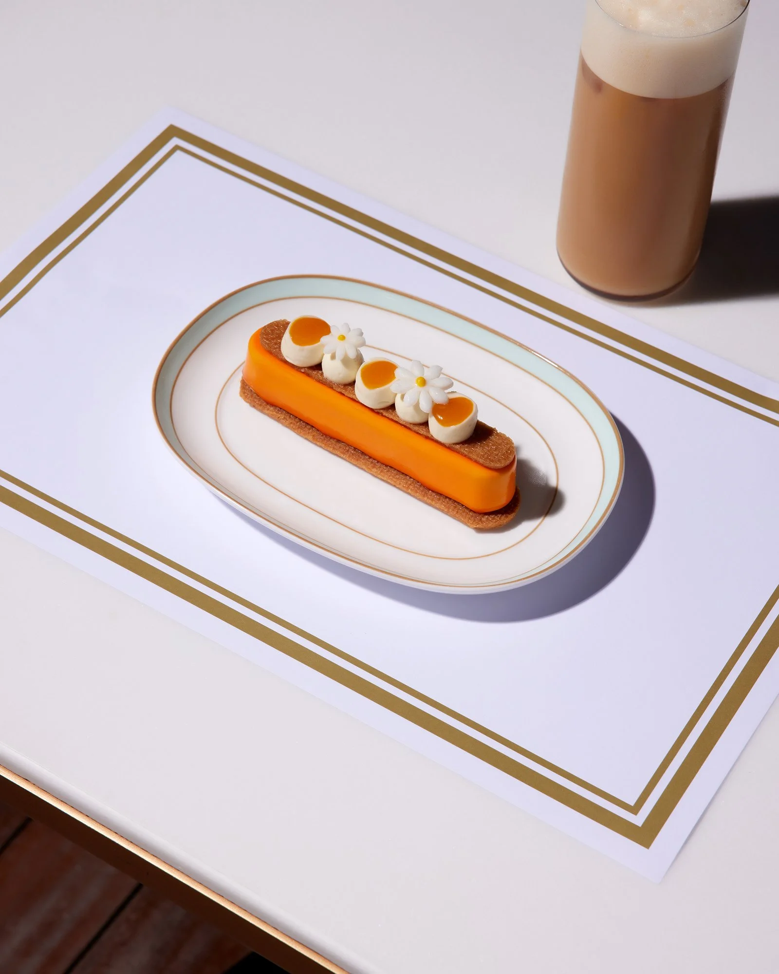 Beyaz bir tabakta turuncu, beyaz ve kahverengi süslemeli bir tatlı; yanında beyaz bir masada buzlu kahve bardağı.