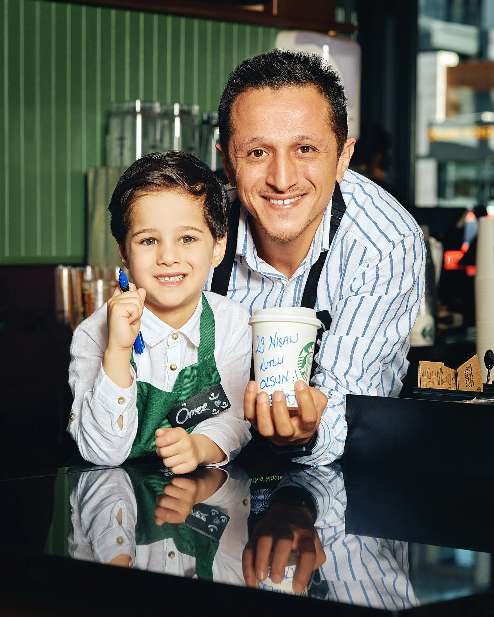 Bir kahve dükkânında, ikisi de önlük giyen bir adam ve bir çocuk. Adam, üzerinde Türkçe el yazısı olan bir Starbucks bardağı tutuyor; çocuk ise elinde bir kalemle gülümseyerek poz veriyor. Çocuğun yaka kartında “Ömer” yazıyor.