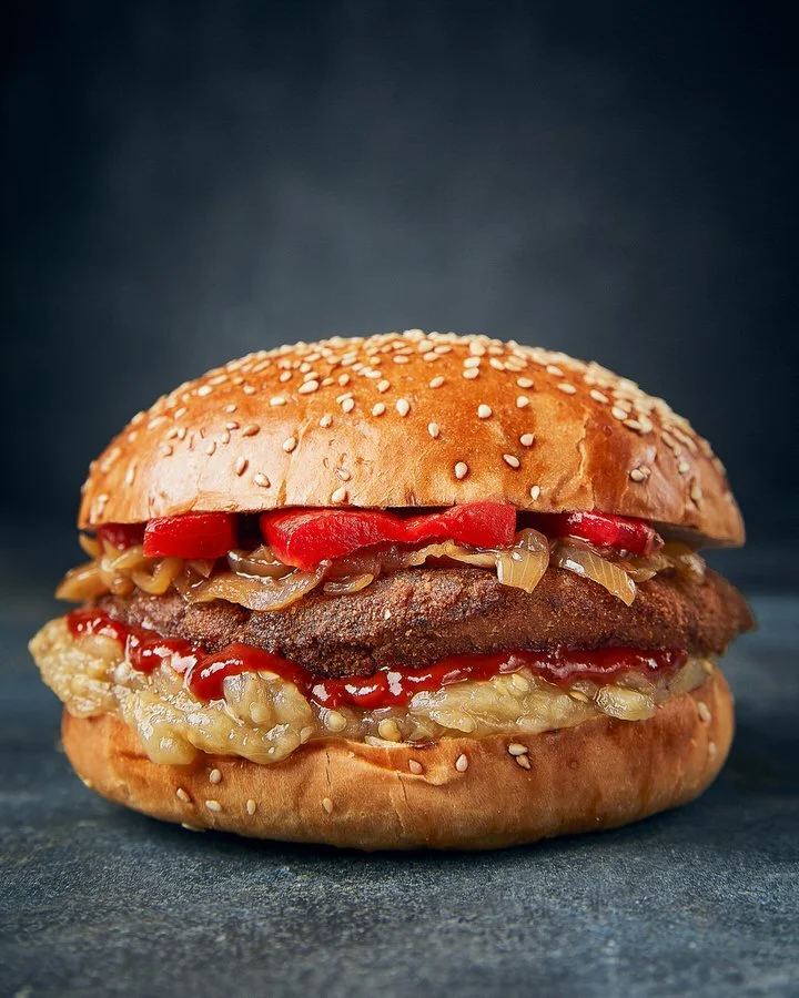 Close-up of a hamburger with a sesame seed bun, beef patty, grilled onions, roasted red peppers, and melted cheese, with ketchup visible, on a dark background.