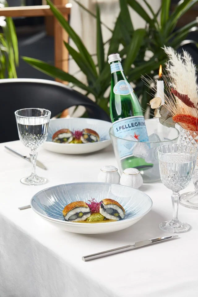 Elegant dining table setting with gourmet dish, S.Pellegrino sparkling water bottle, crystal glasses, and decorative plants.