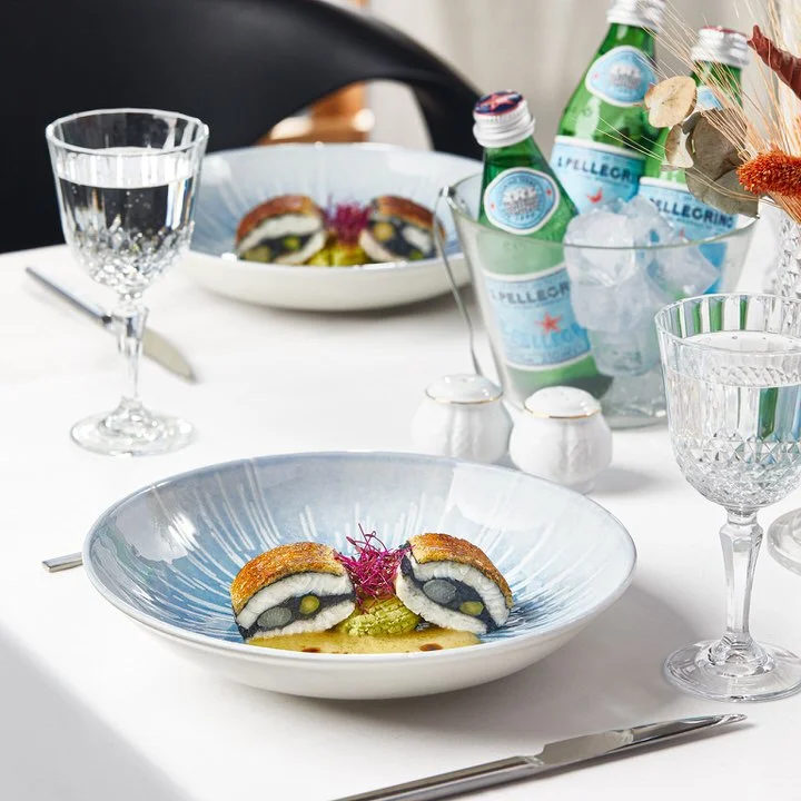 Elegant dining table setup with two plates featuring a gourmet dish. Each plate contains a carefully arranged culinary creation, possibly a stuffed roll with sauce and garnishes, on a white tablecloth. Crystal glasses are filled with water, and there