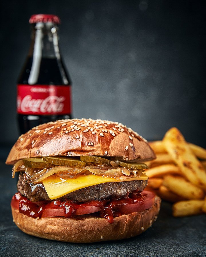 Close-up of a cheeseburger with lettuce, tomato, pickles, and onions, accompanied by fries and a bottle of Coca-Cola in the background on a dark surface.