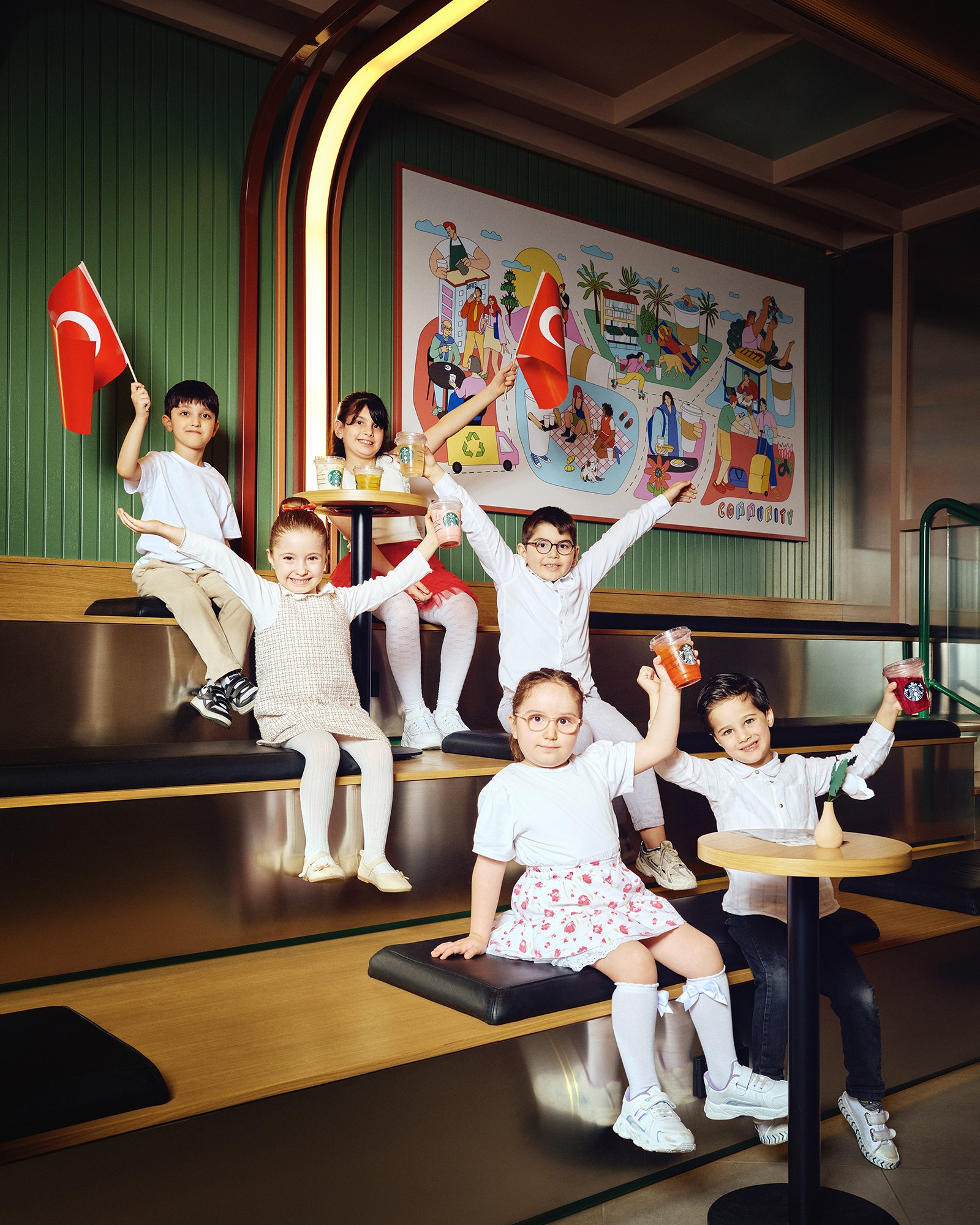 Modern bir iç mekânda, Türk bayraklarıyla kutlama yapan yedi çocukluk bir grup; arka planda topluluk temalı renkli bir duvar resmi bulunuyor.