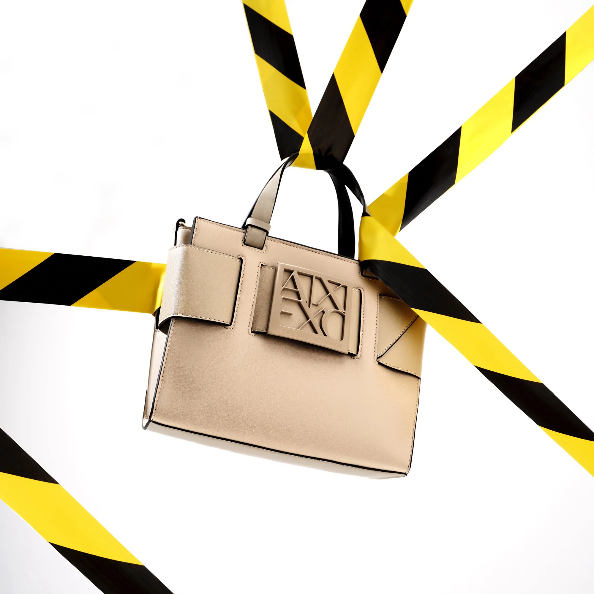 Belirgin logo etiketine sahip bej bir tasarımcı çantası, beyaz bir arka plan üzerinde sarı-siyah uyarı bantlarıyla çevrilmiş şekilde duruyor.