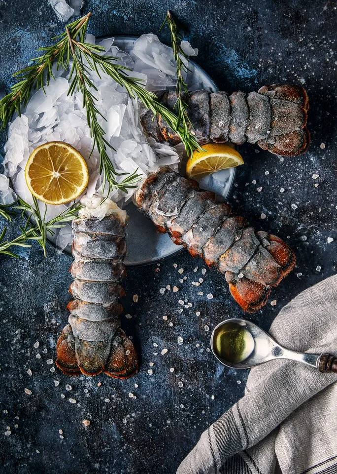 Buz üzerinde servis edilen üç dondurulmuş ıstakoz kuyruğu, biberiye ve limon dilimleriyle süslenmiş, yanında tuz serpilmiş ve sos kaşığı yer alıyor.