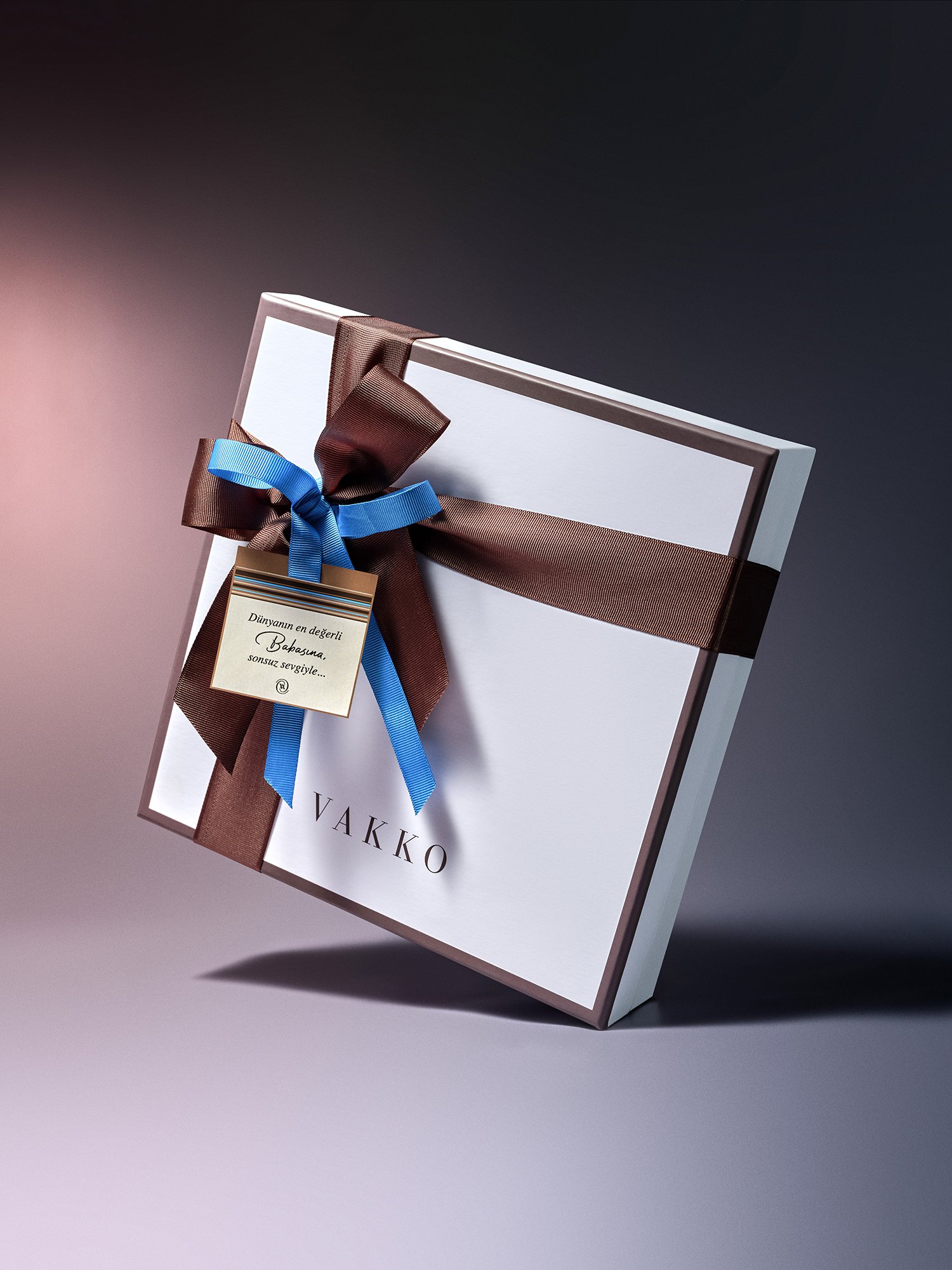 Kahverengi kurdele ve mavi fiyonkla sarılmış, etiket takılı bir hediye kutusu; gradyan bir arka plan önünde duruyor.