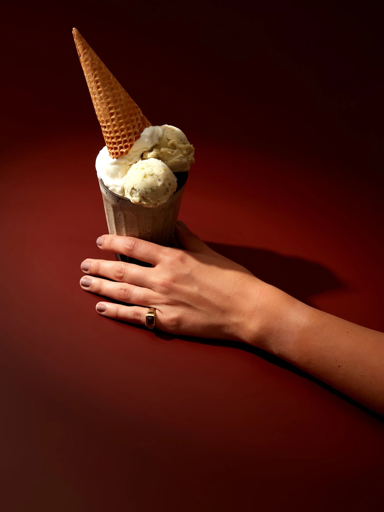 Kırmızı bir yüzey üzerinde duran, parmağında altın yüzük takılı bir el; üstünde külah gofreti bulunan vanilyalı dondurma bardağı tutuyor.