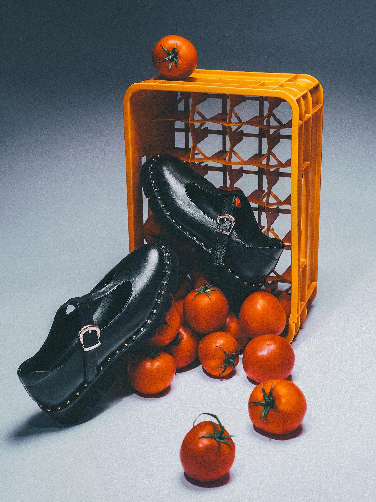 Turuncu plastik bir kasadan dökülen turuncu domateslerin arasında, zımbalı siyah ayakkabılar yer alıyor.