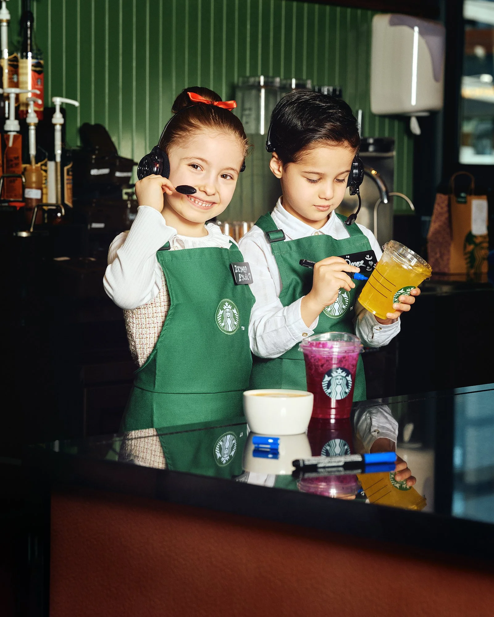 Starbucks’ta barista olarak çalışan iki küçük çocuk; yeşil önlükler ve kulaklıklı mikrofon takıyorlar, önlerinde içecekler duruyor.