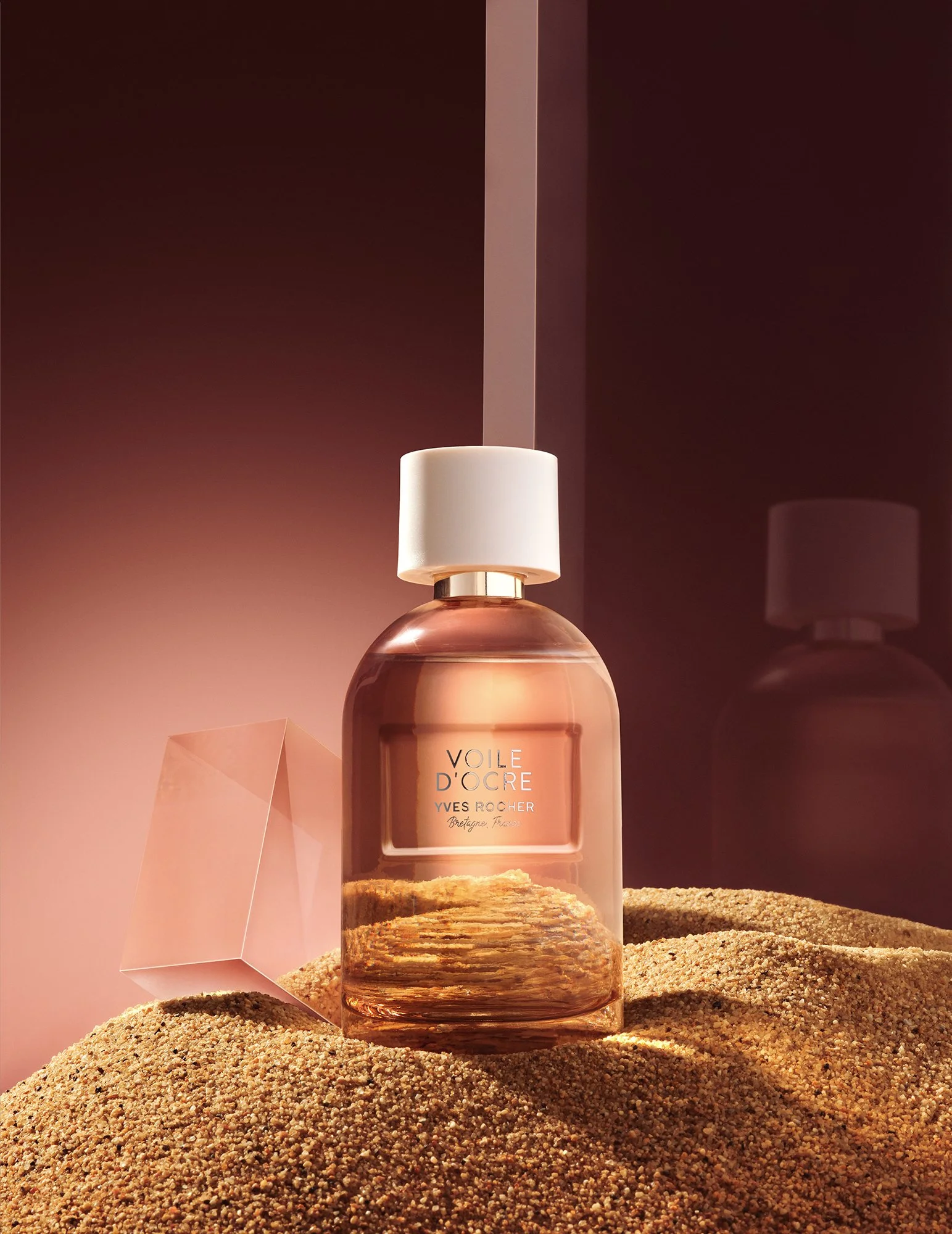 “Voile D’Ocre Yves Rocher” etiketli bir parfüm şişesi; kumlu bir yüzey üzerinde, yanında pembe geometrik bir obje ile duruyor. Arka plan yansıtıcı pembe bir duvar.