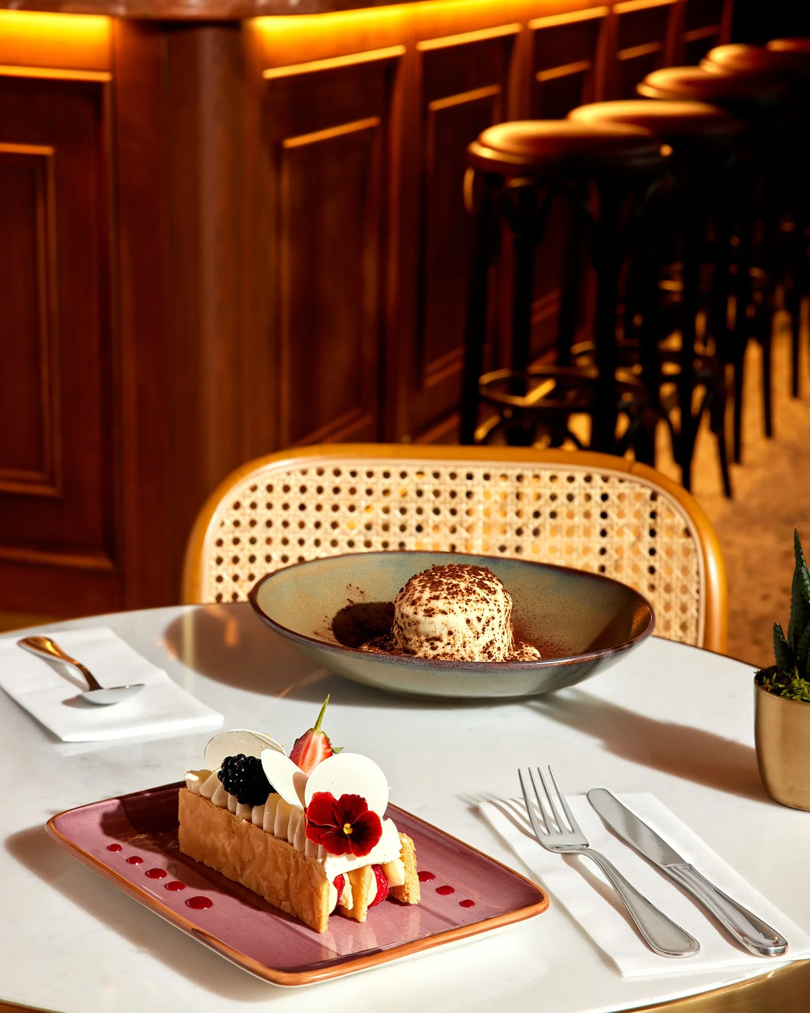 Ahşap panelli ve bar tabureli şık bir restoranda; üzeri meyveler ve yenilebilir çiçeklerle süslenmiş dikdörtgen bir tatlının bulunduğu masa, yanında krema ve kakao tozu içeren yuvarlak bir kahve/tatlı tabağıyla birlikte.