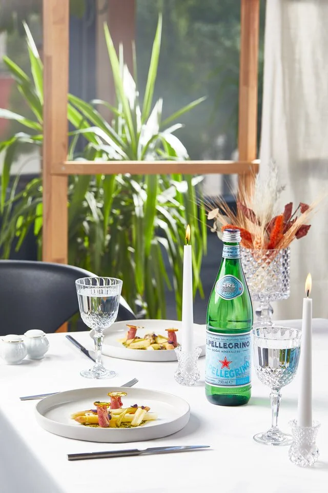 Şık bir yemek masası düzeni, gurme yemek tabakları, su bardakları, mumlar ve dekoratif bitkilerle süslenmiş.

