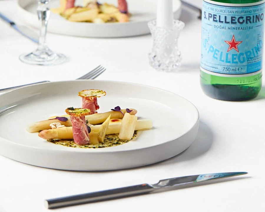 Beyaz tabakta kuşkonmaz ve prosciutto ile süslenmiş gurme yemek, yanında bir şişe maden suyu.

