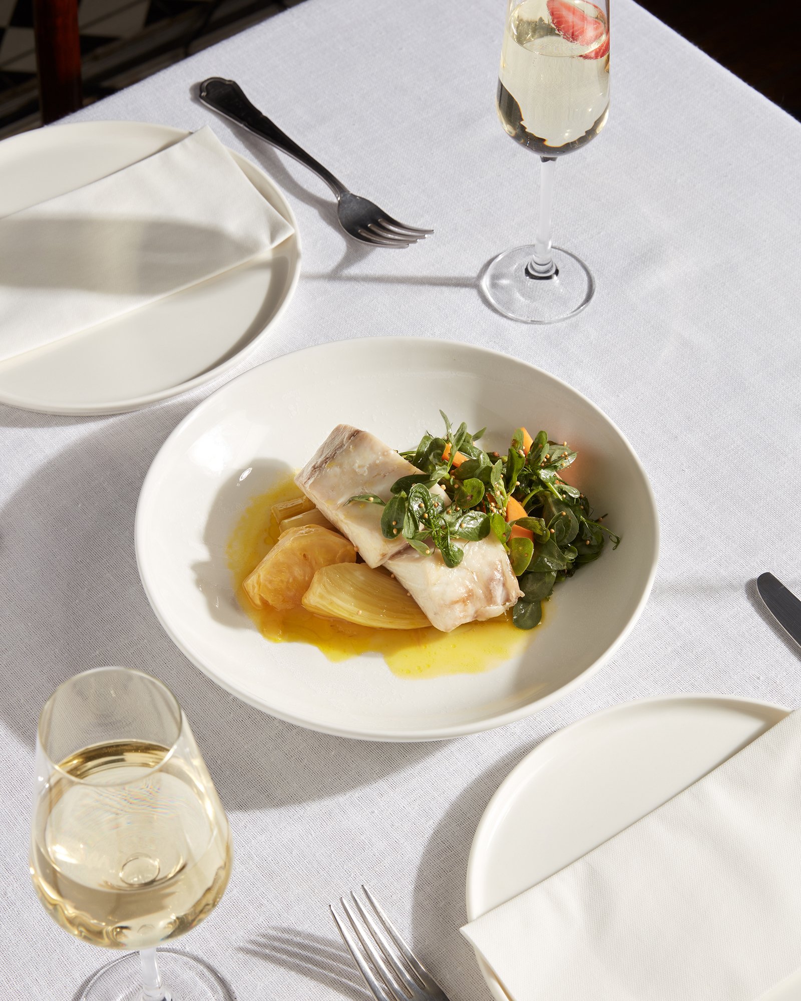 Beyaz örtülü, özenle hazırlanmış bir yemek masası; sebze ve sos eşliğinde balık ana yemek tabağı, iki kadeh beyaz şarap ve boş tabaklarla çatal-bıçak takımları yer alıyor.