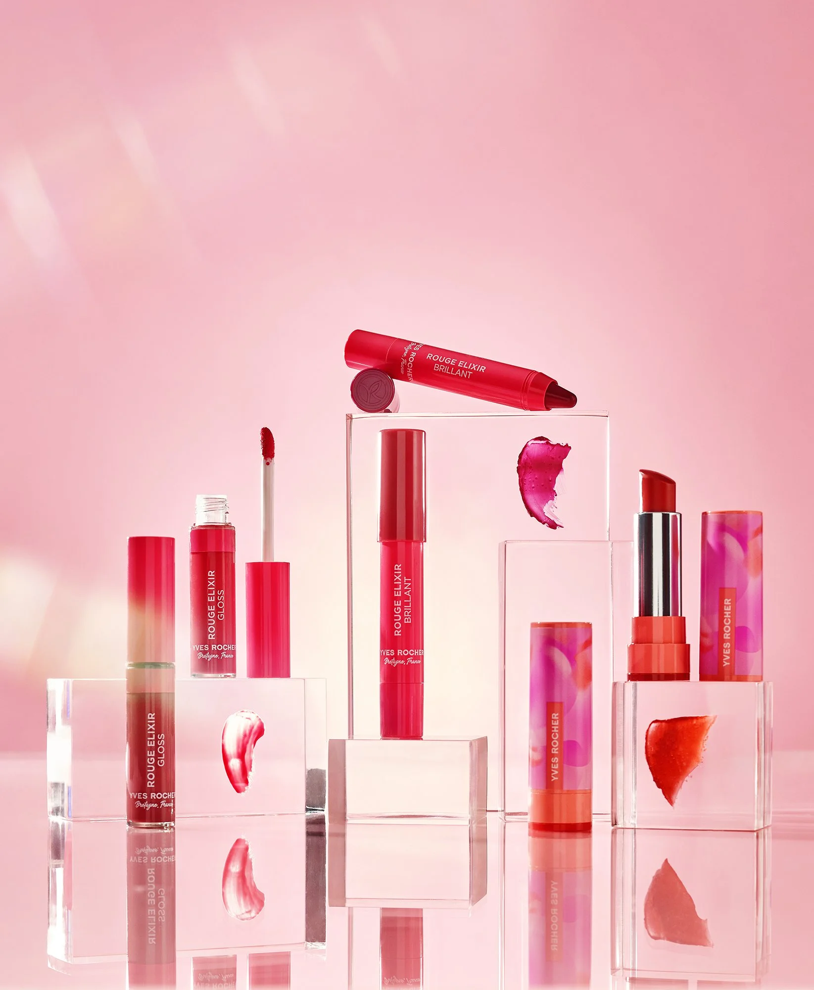 Multiple shades of red and pink lipstick and lip gloss tubes displayed on clear acrylic blocks, with some lipstick swatches and a pink background.