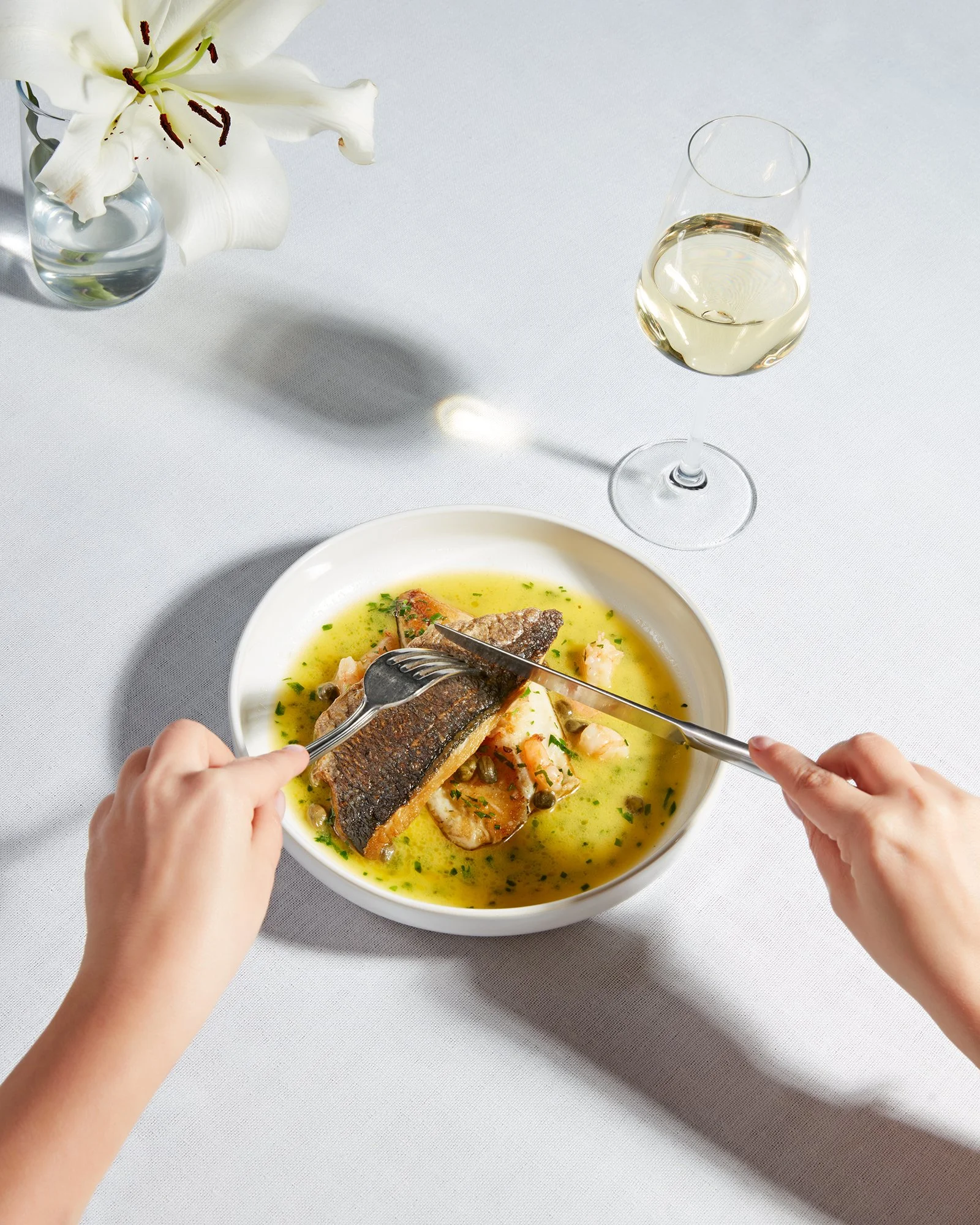 Otlarla hazırlanmış balık çorbasının içindeki bir balık parçasını kesen eller; yanında bir kadeh beyaz şarap ve beyaz zambak bulunan bir vazo, hepsi beyaz örtülü bir masada.