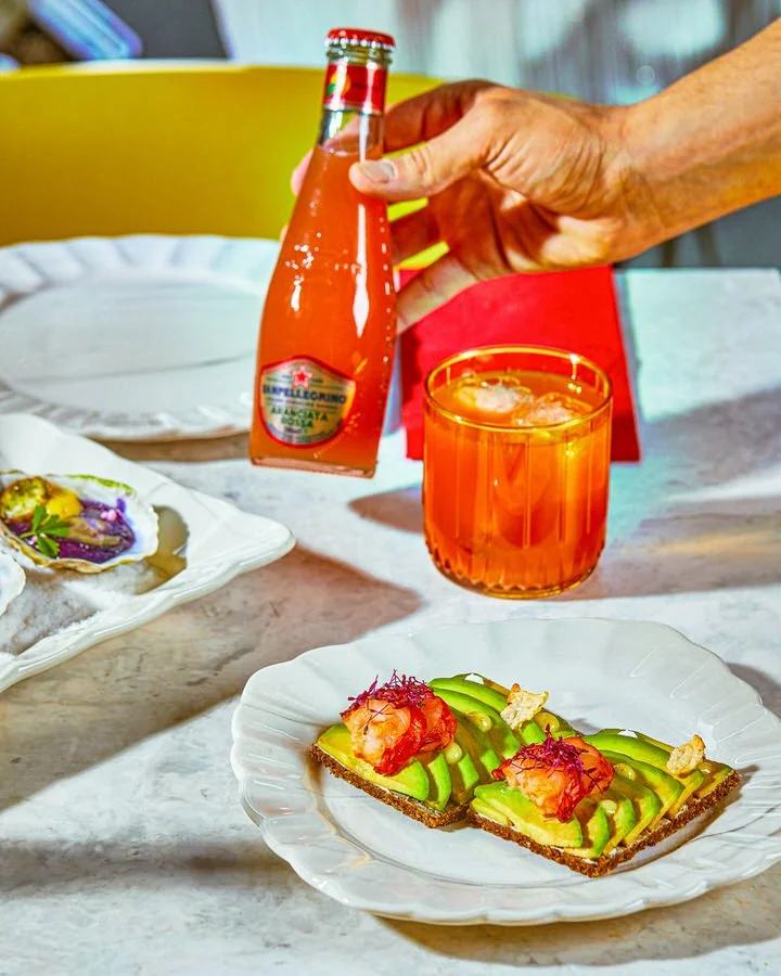 Bir kişi, bir şişe gazlı içecek tutuyor, yanında portakal içeceği bardağı ve avokado tost bulunan masa düzeni.

