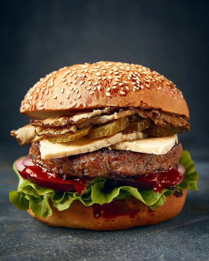 Gourmet burger with sesame seed bun, beef patty, lettuce, tomato, pickles, cheese, mushrooms, and ketchup.